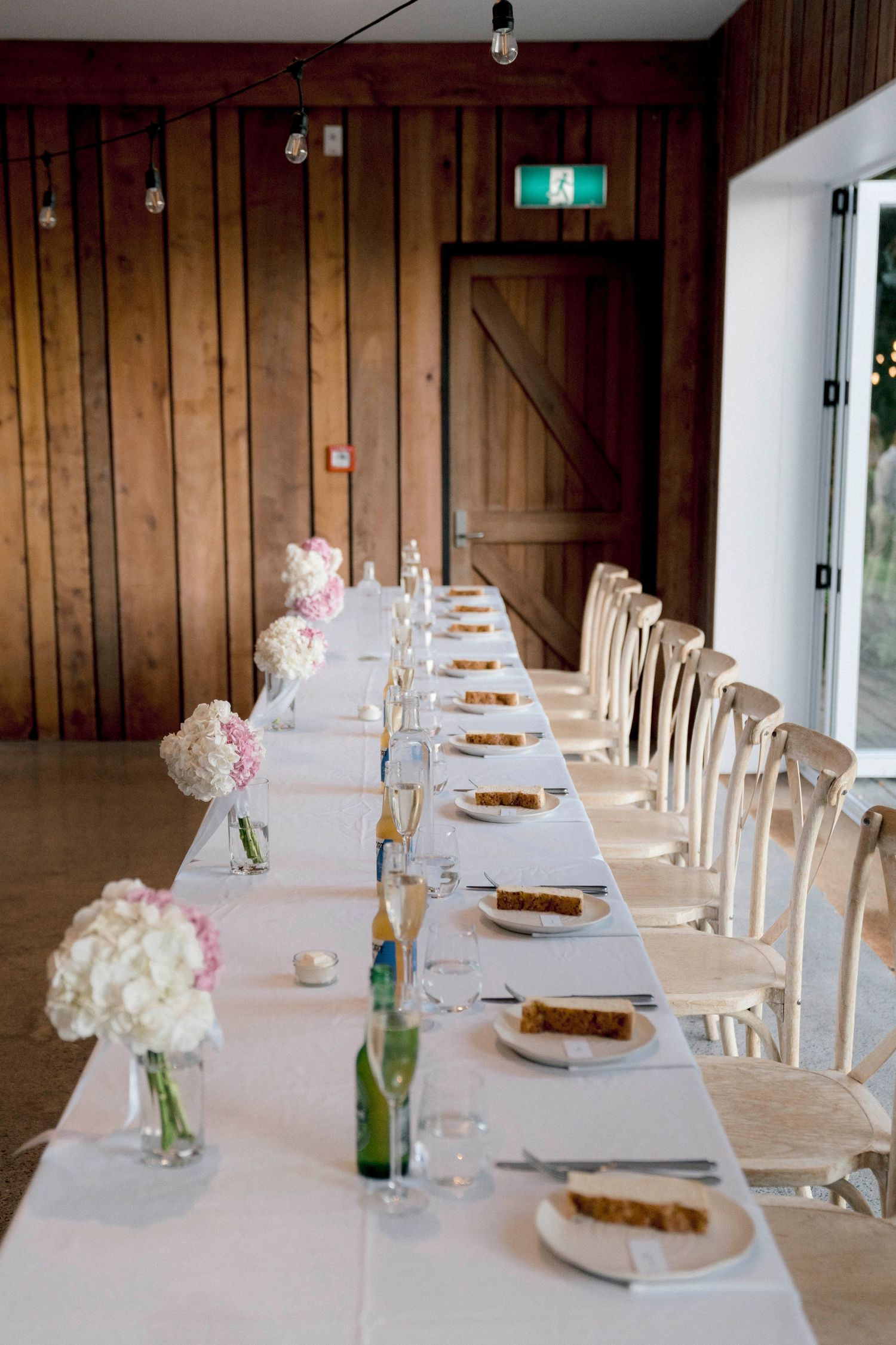 Long banquet table set with white linens, glass bottles and floral centerpieces against rustic wood-paneled walls.