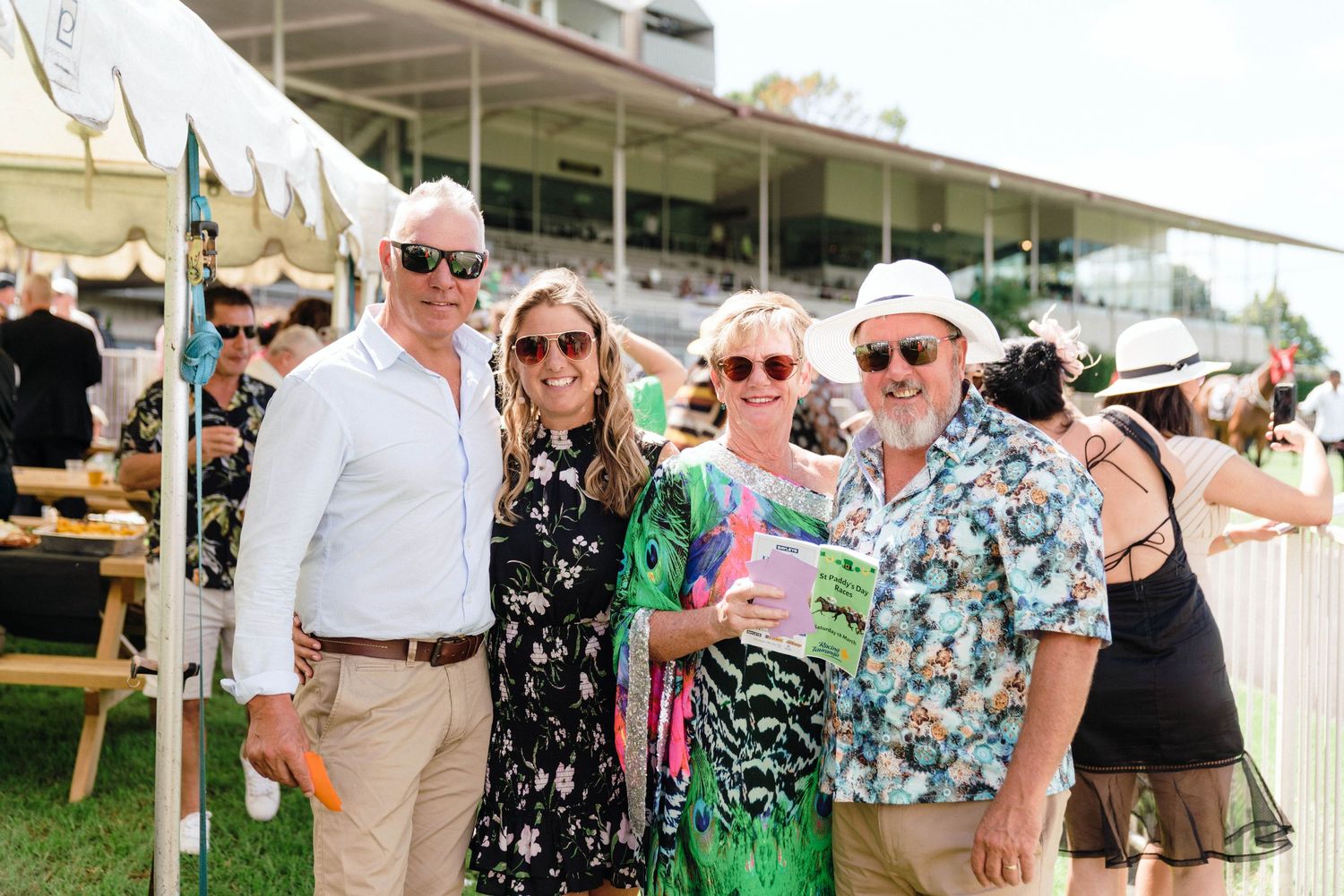 Guests in summer attire gathered at an outdoor event with tents and umbrellas in the background.