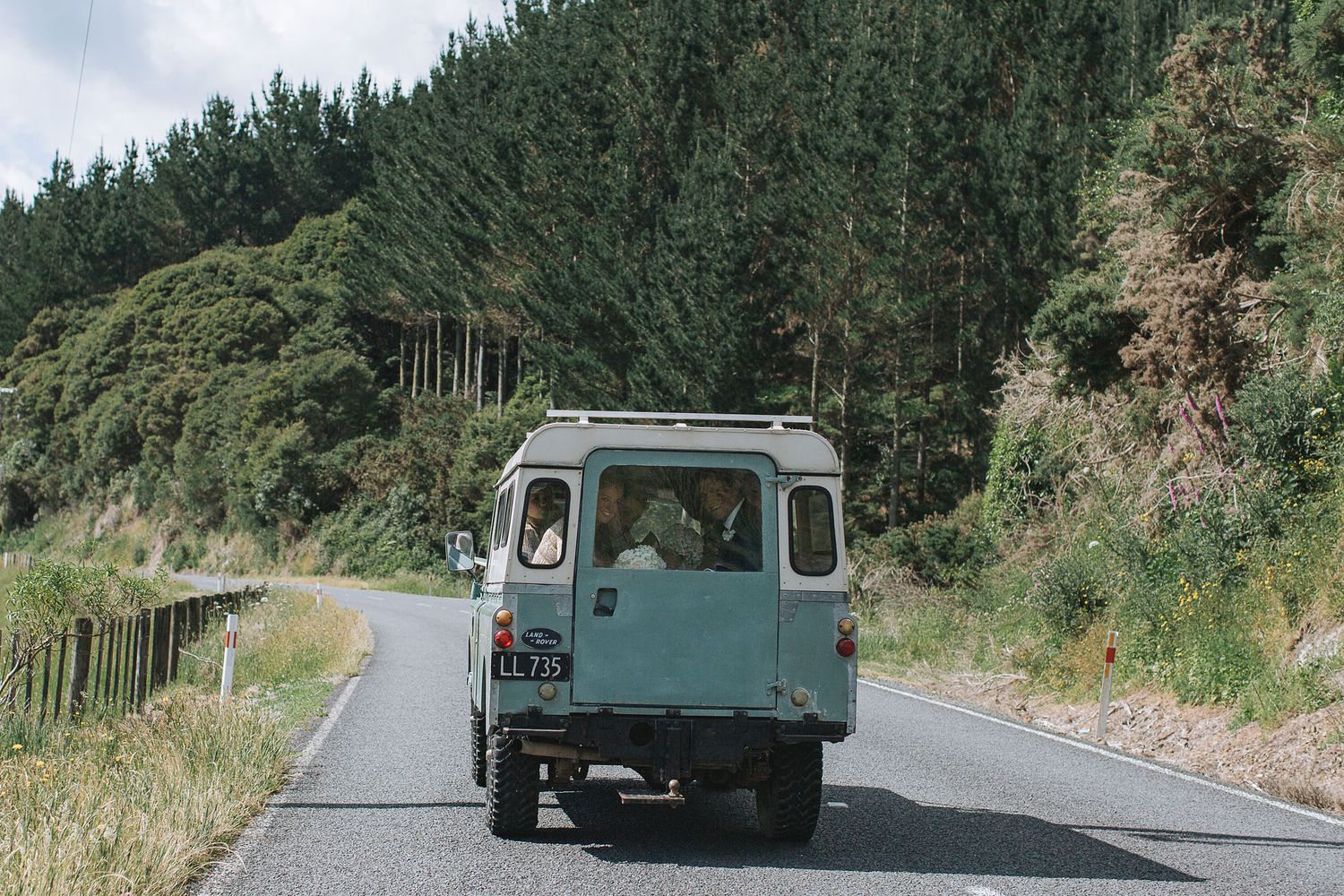A vintage blue van drives down a winding mountain road surrounded by pine trees and green hills.