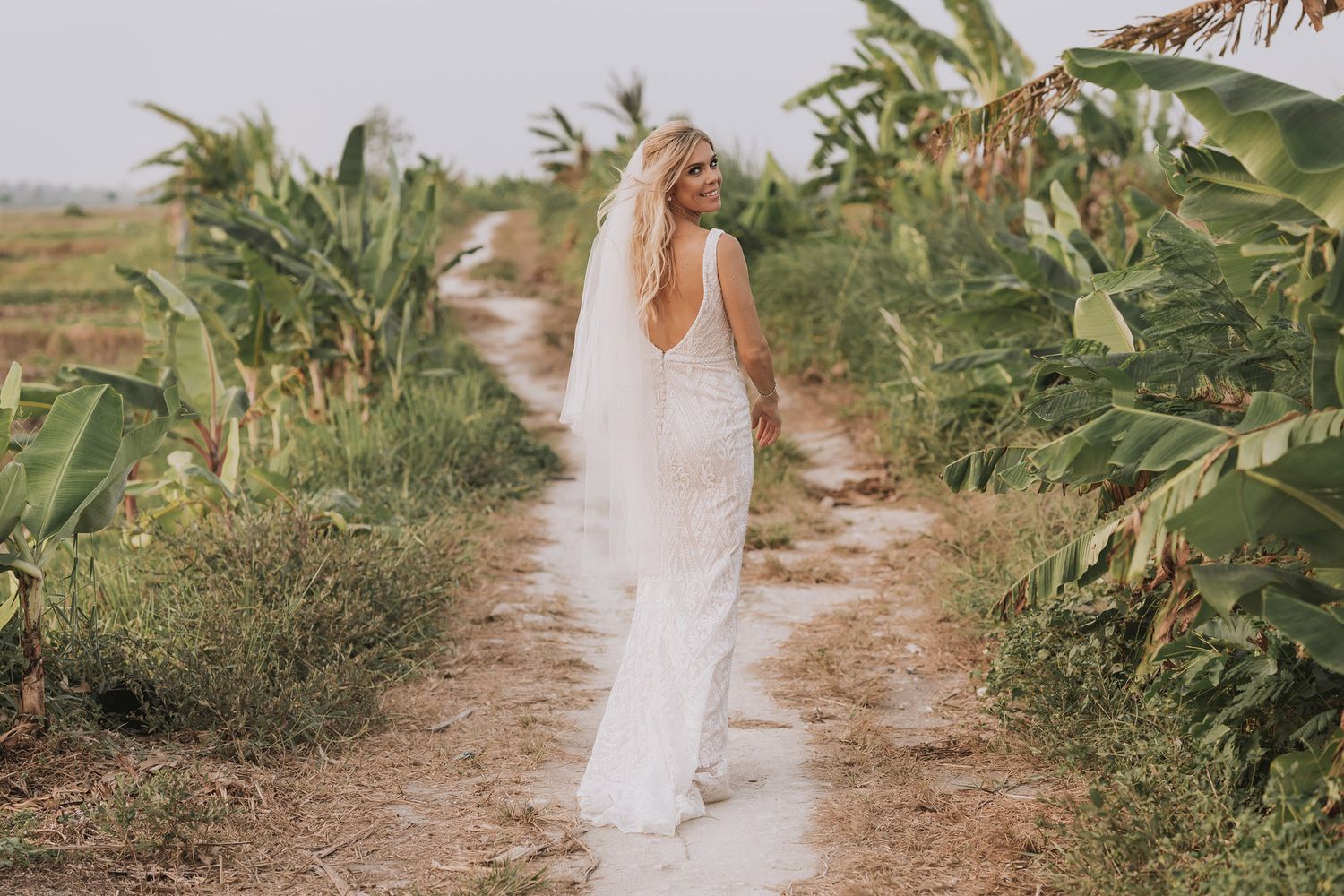 Person in flowing white dress walking along dirt path bordered by tropical foliage.
