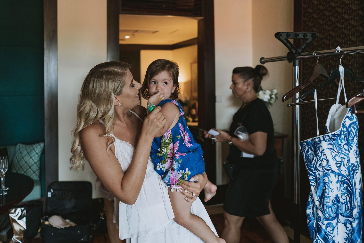 A person in a white dress holds a small child in a floral dress while getting ready in a hotel room.