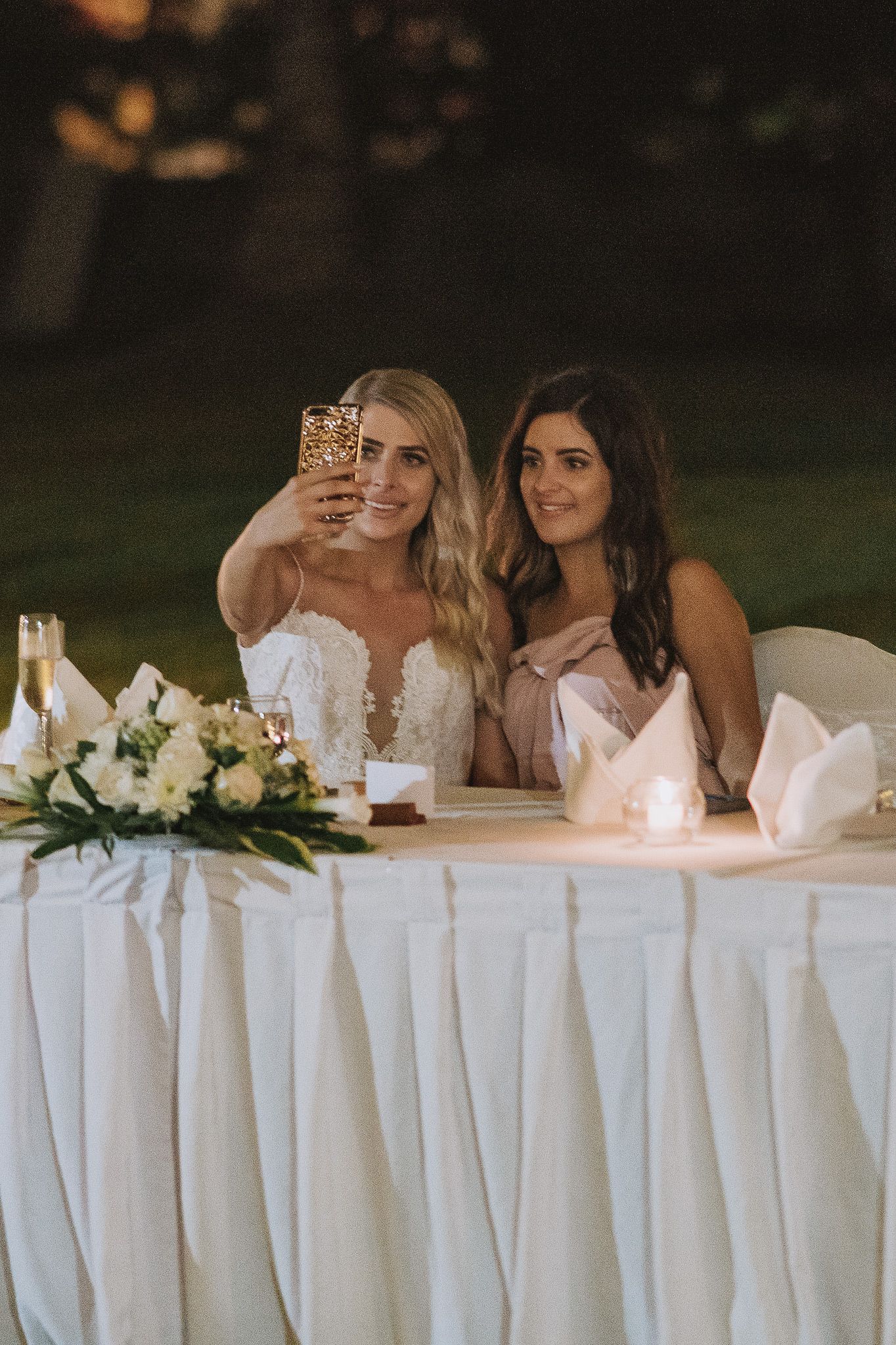 Two women in formal attire taking a selfie at an elegant reception table with white floral centerpieces.