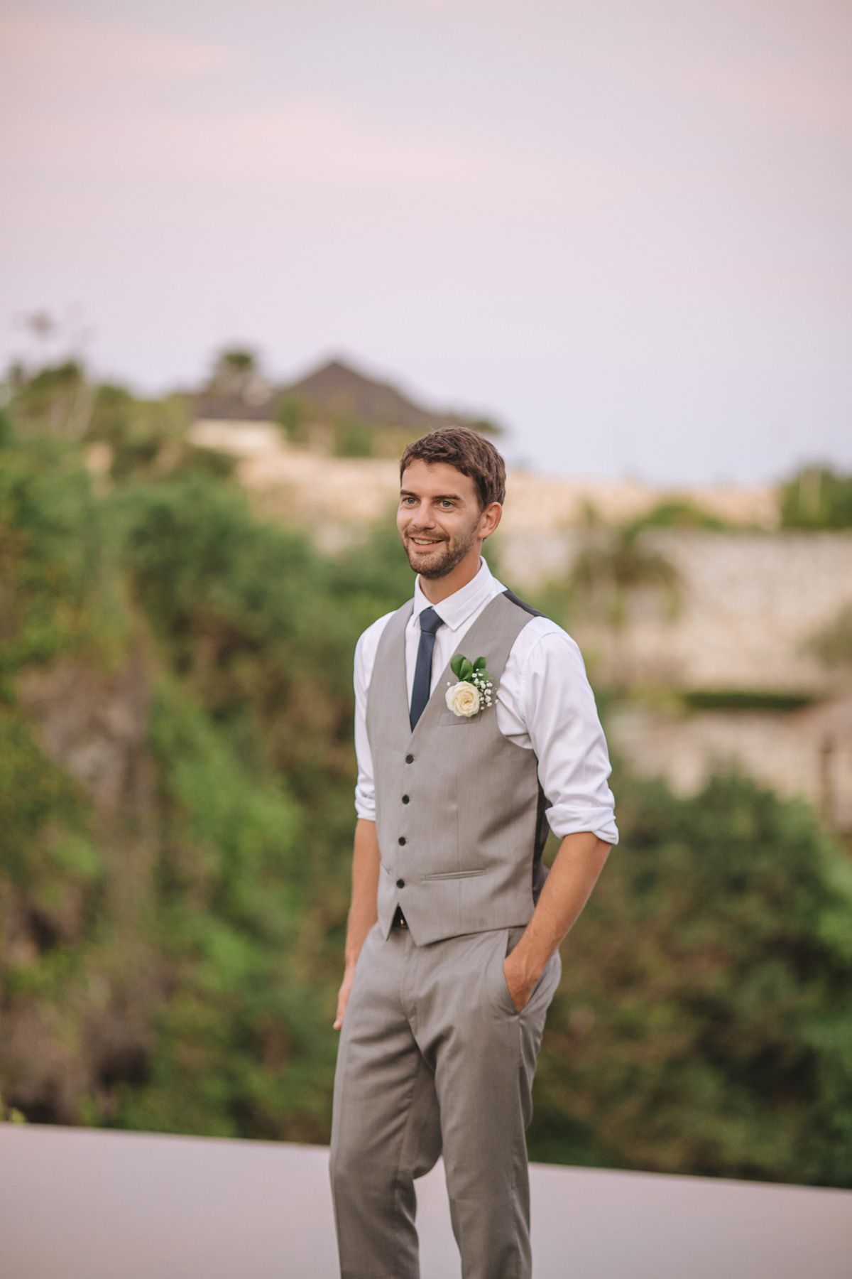 A groom in a gray vest and white shirt poses for portraits in an outdoor garden setting.
