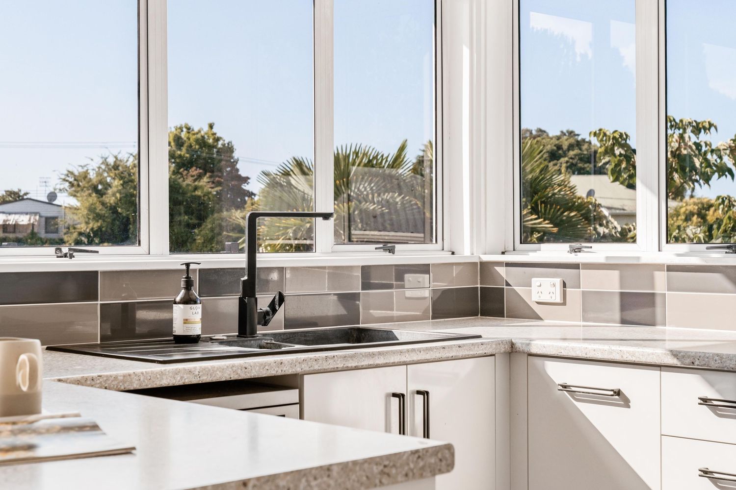 Modern white kitchen with granite countertops and large windows overlooking green outdoor scenery.