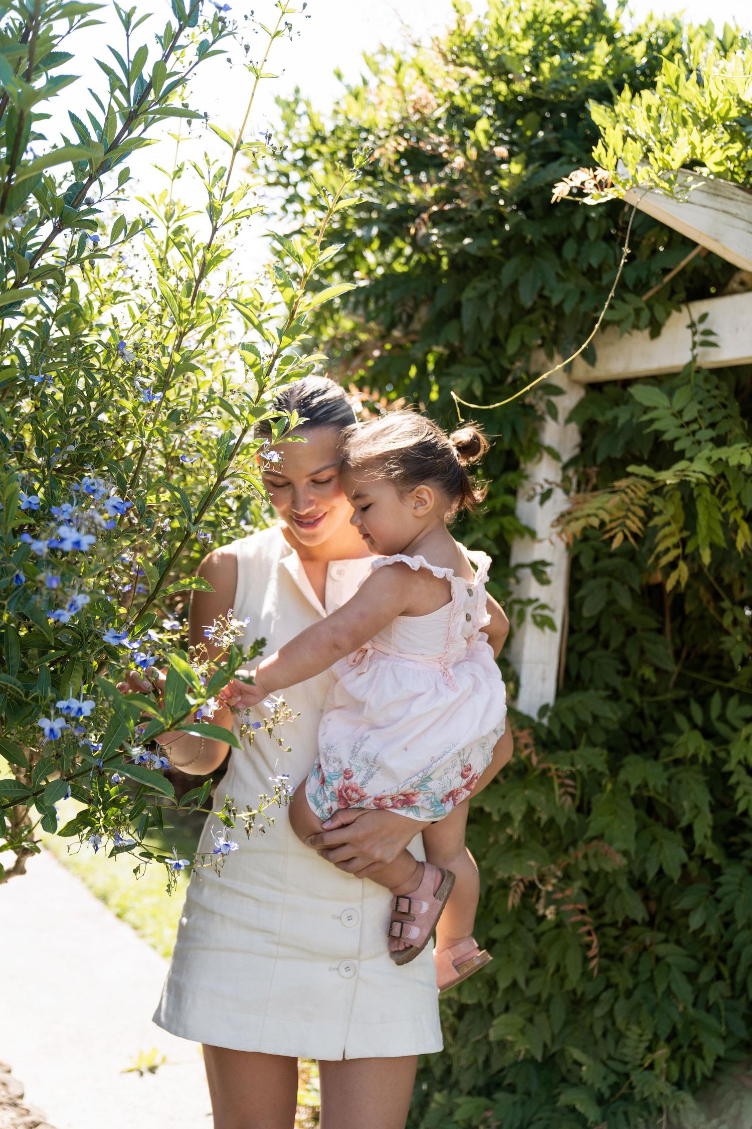 Two figures in white dresses share tender moments in a sunny garden setting with greenery in the background.
