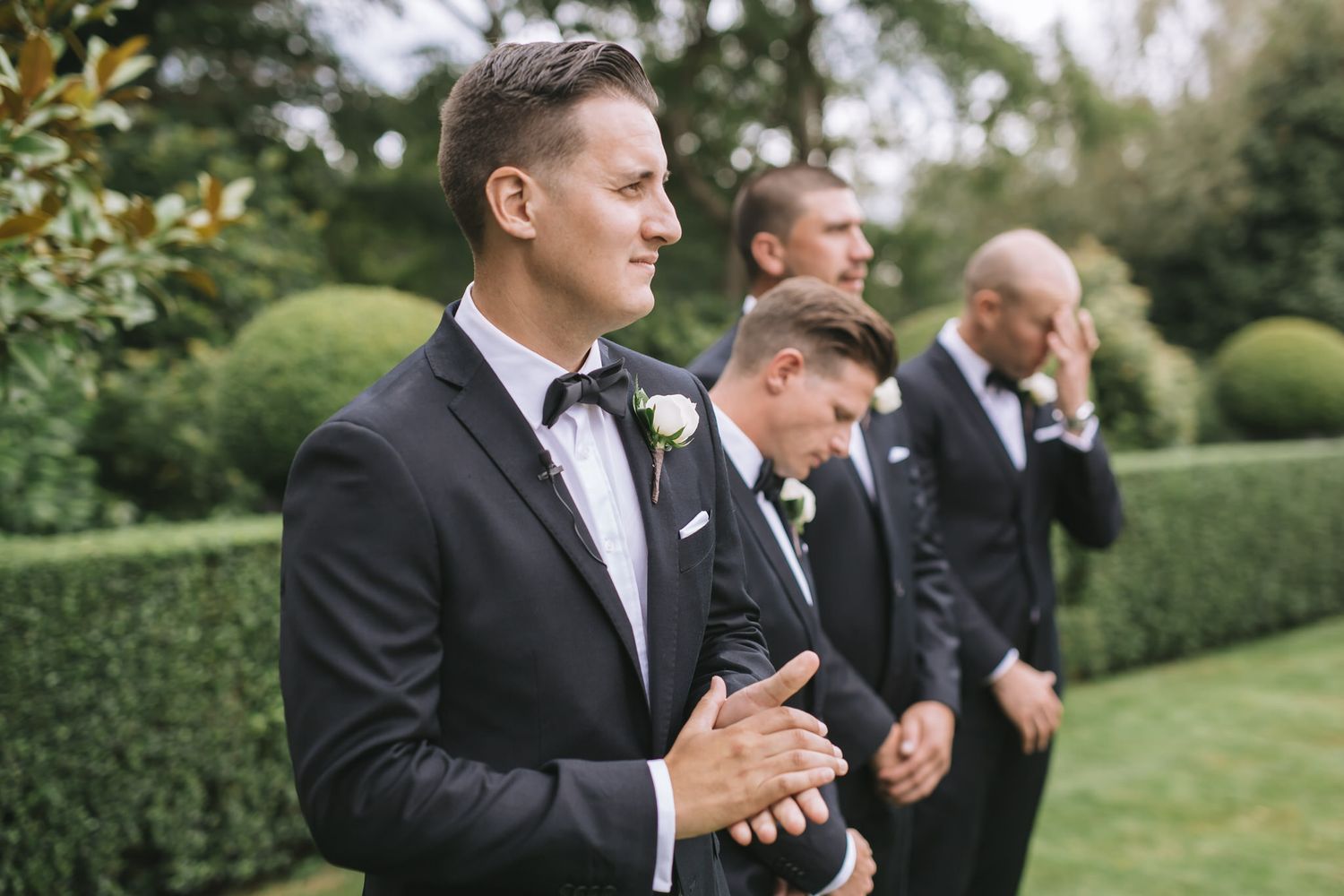 Groomsmen in classic black tuxedos line up on green lawn against manicured hedge backdrop at outdoor wedding ceremony.
