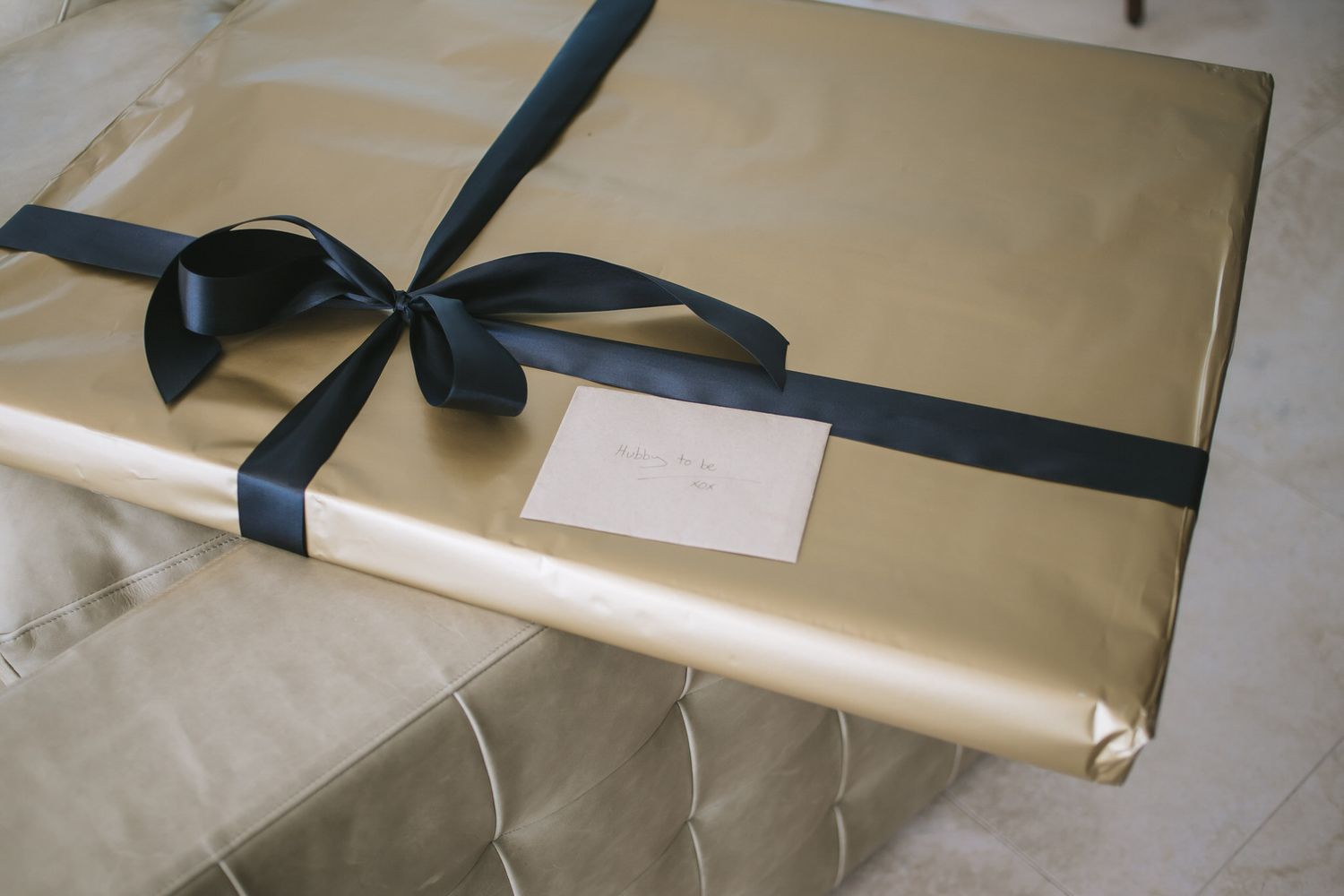 A gold-wrapped present box tied with an elegant black satin ribbon and small white gift tag on gray marble surface.