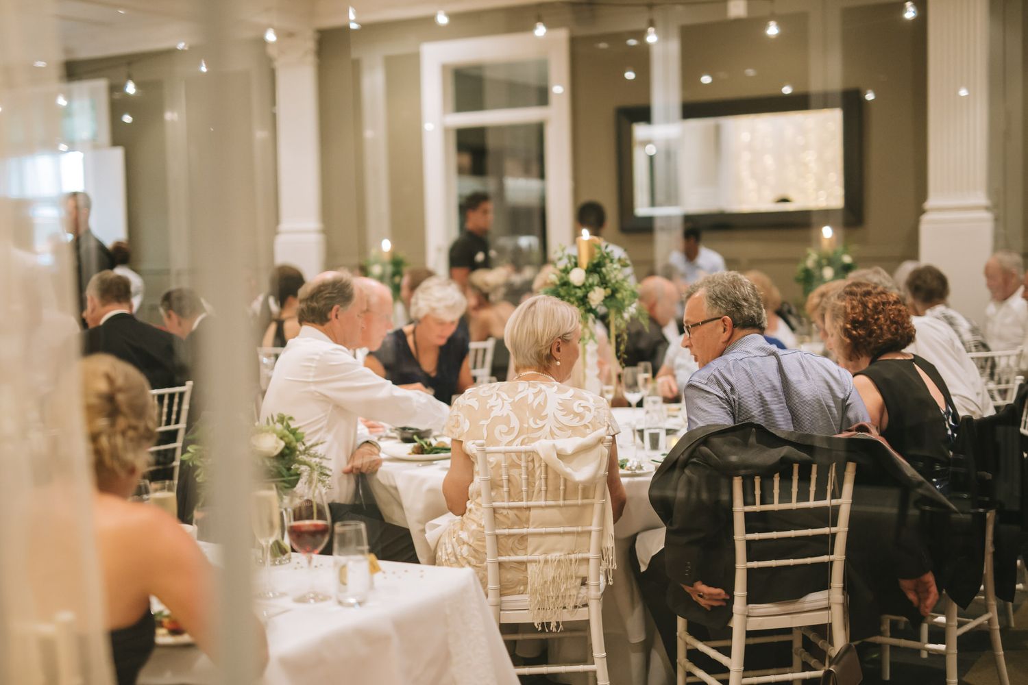 Guests seated at elegant round tables with white linens and chiavari chairs during an indoor wedding reception.