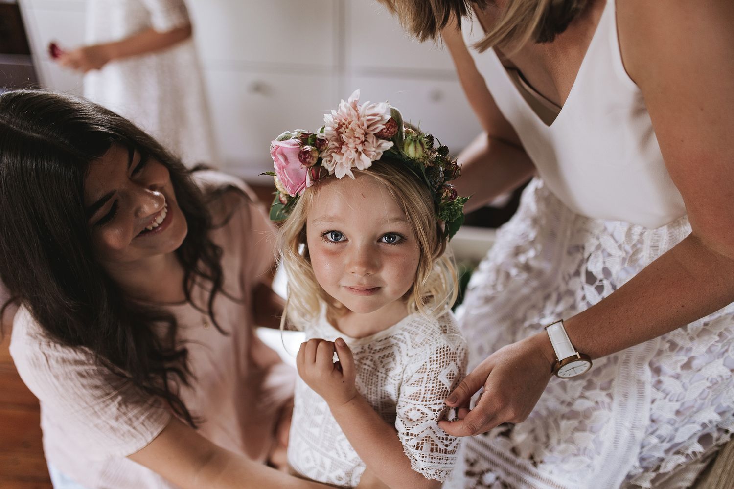 Little girl wearing a floral crown surrounded by people in lace dresses at a wedding preparation.