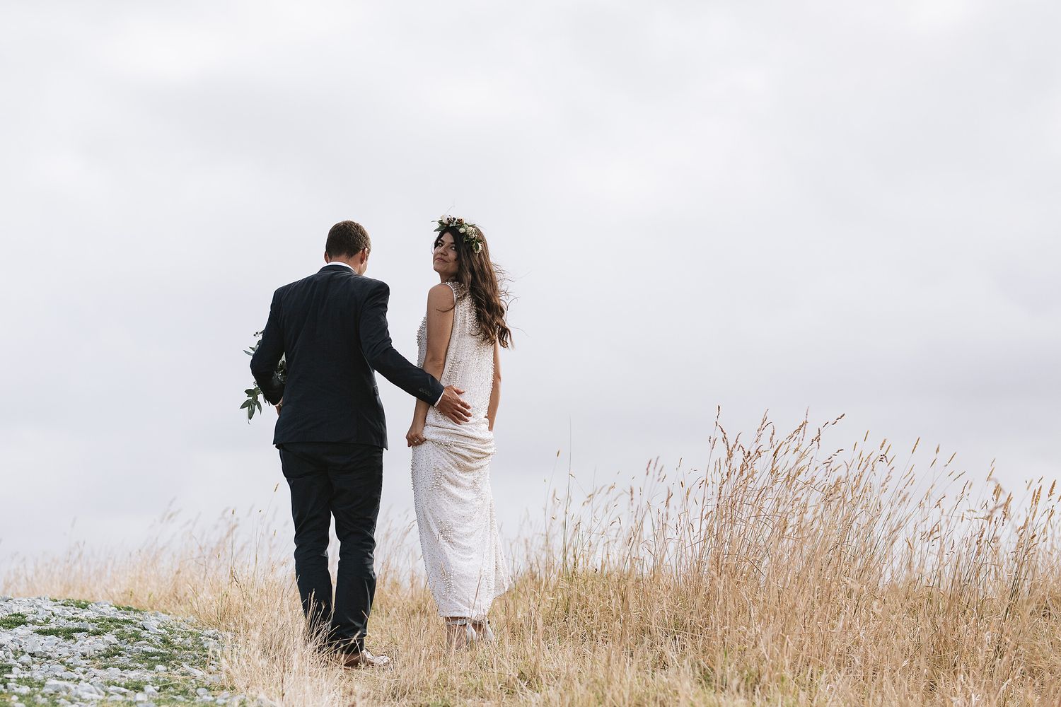 A couple in wedding attire walks through tall grass on a hillside against an overcast sky.