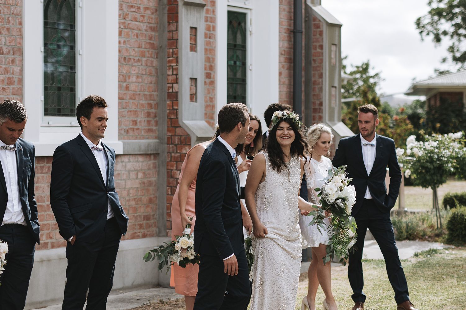 Wedding party in formal attire stand together outside rustic brick building on sunny afternoon.