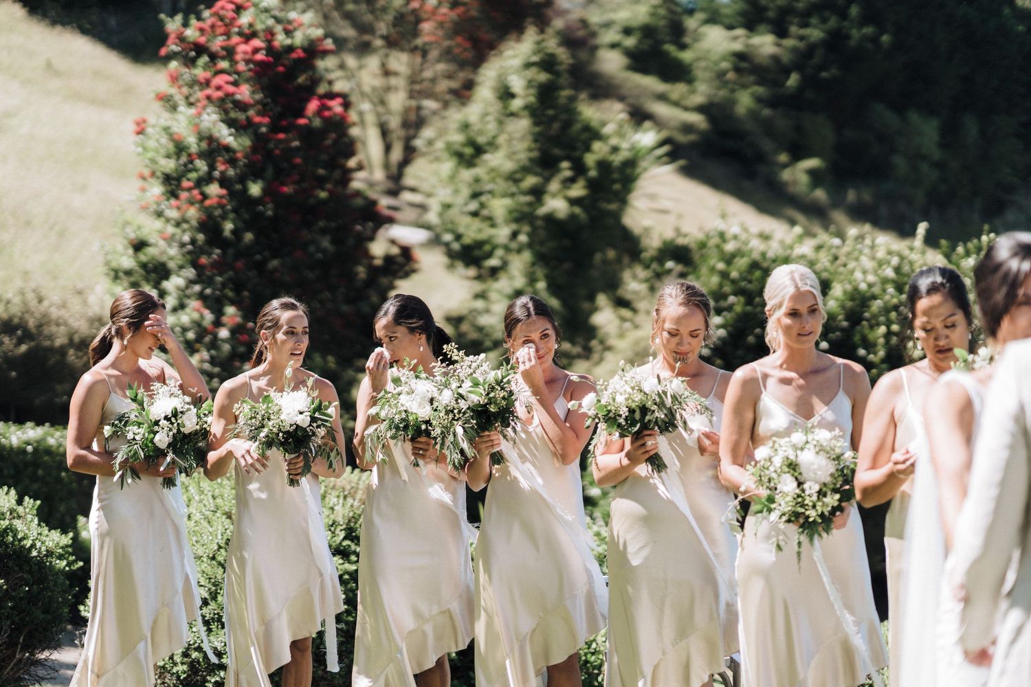 Bridesmaids in cream-colored dresses hold white floral bouquets while standing outdoors on a sunny hillside.