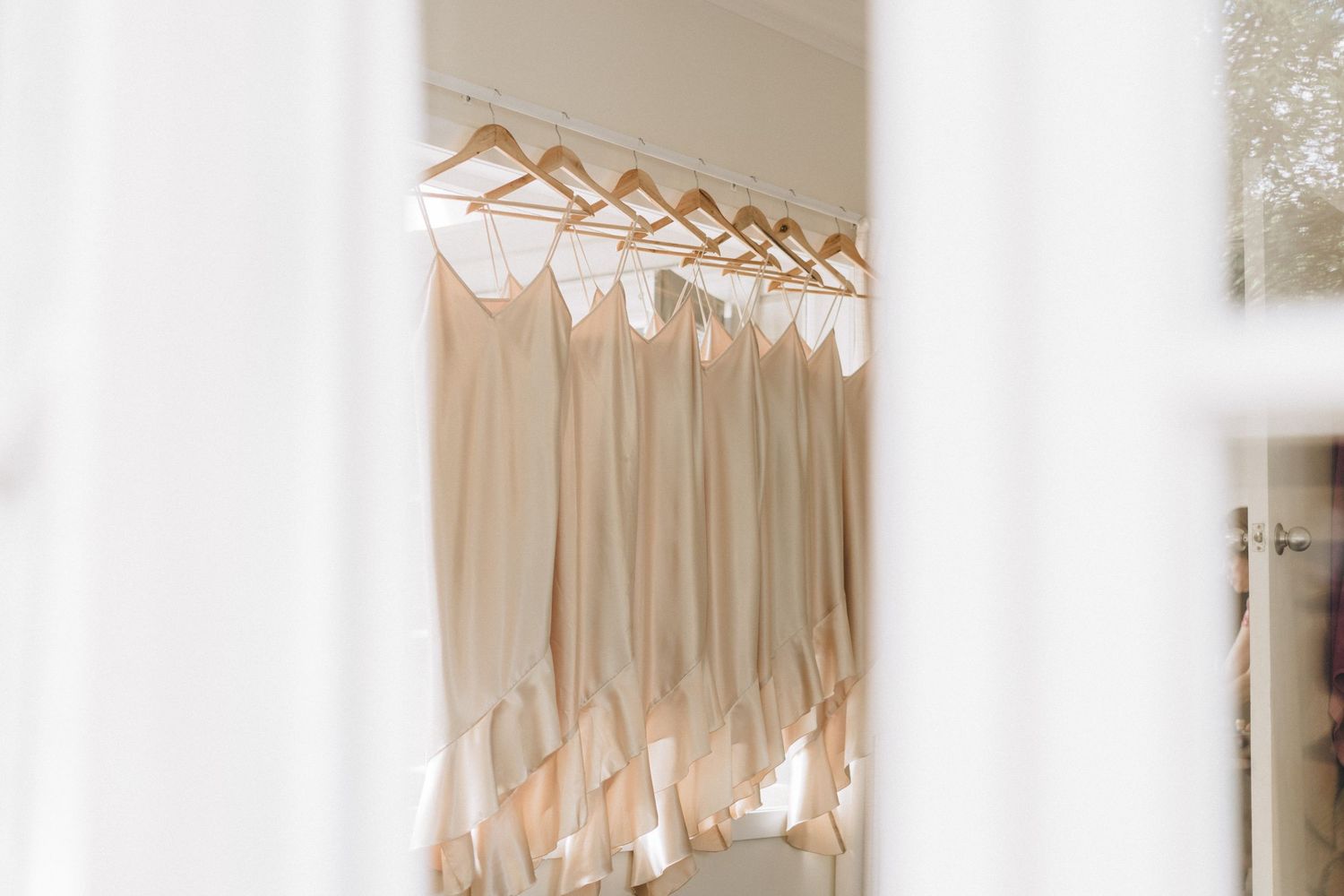 A row of elegant champagne-colored bridesmaid dresses hanging on wooden hangers in a brightly lit closet.