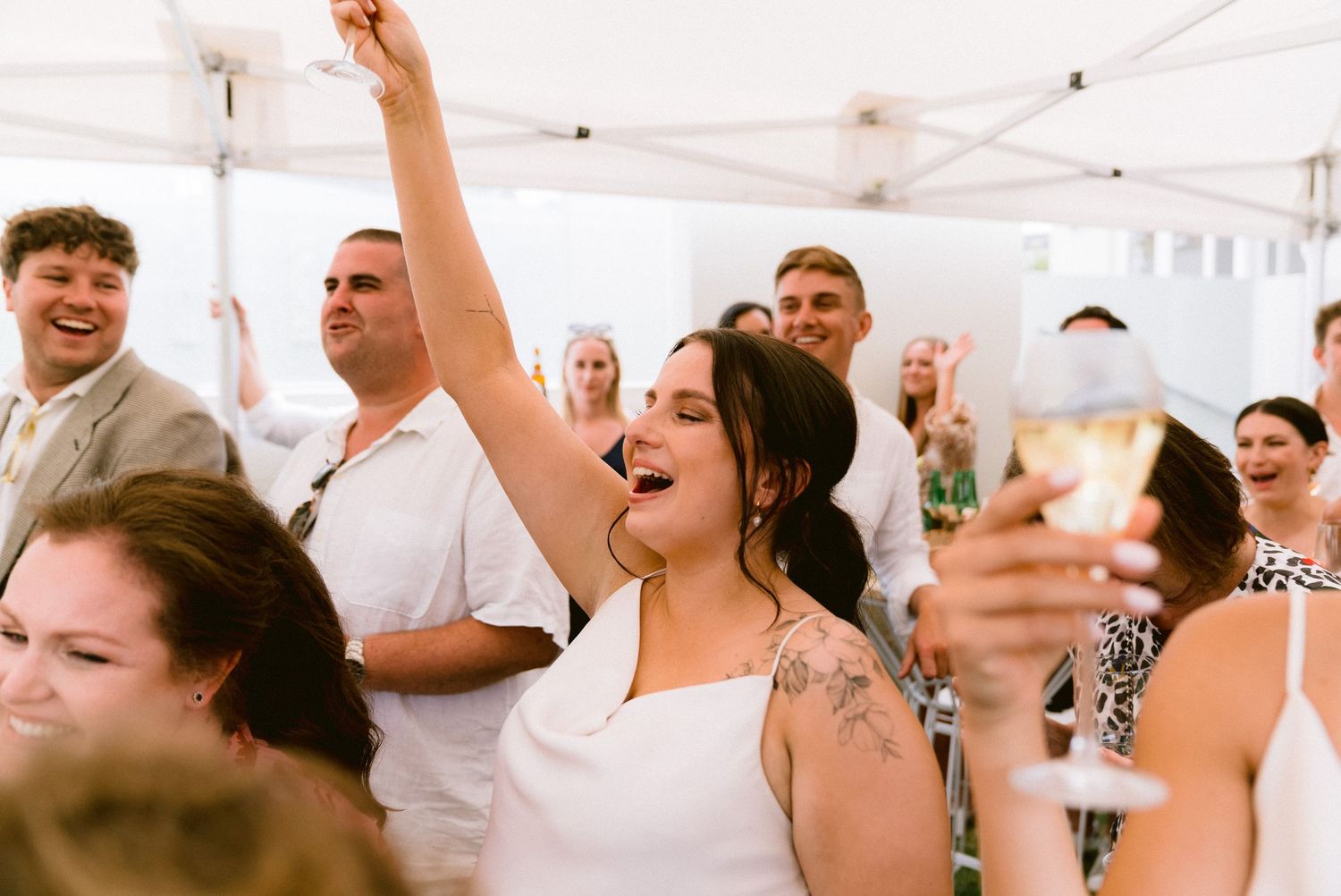 Wedding reception party guests raise their glasses in celebration under a white tent.
