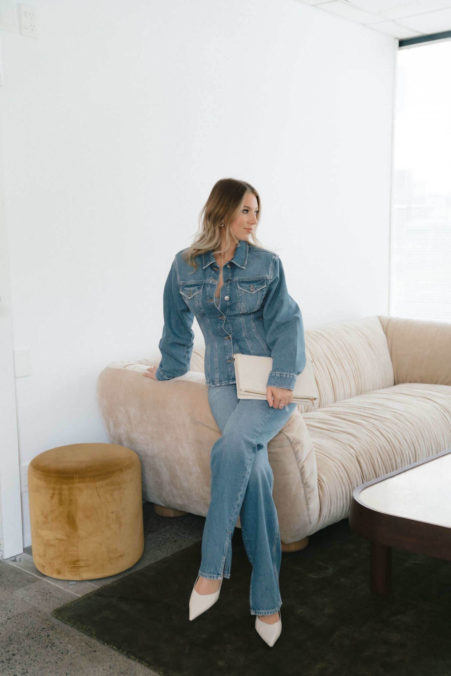 Someone in a denim jacket sits thoughtfully on a beige couch with a golden ottoman nearby in a bright room.