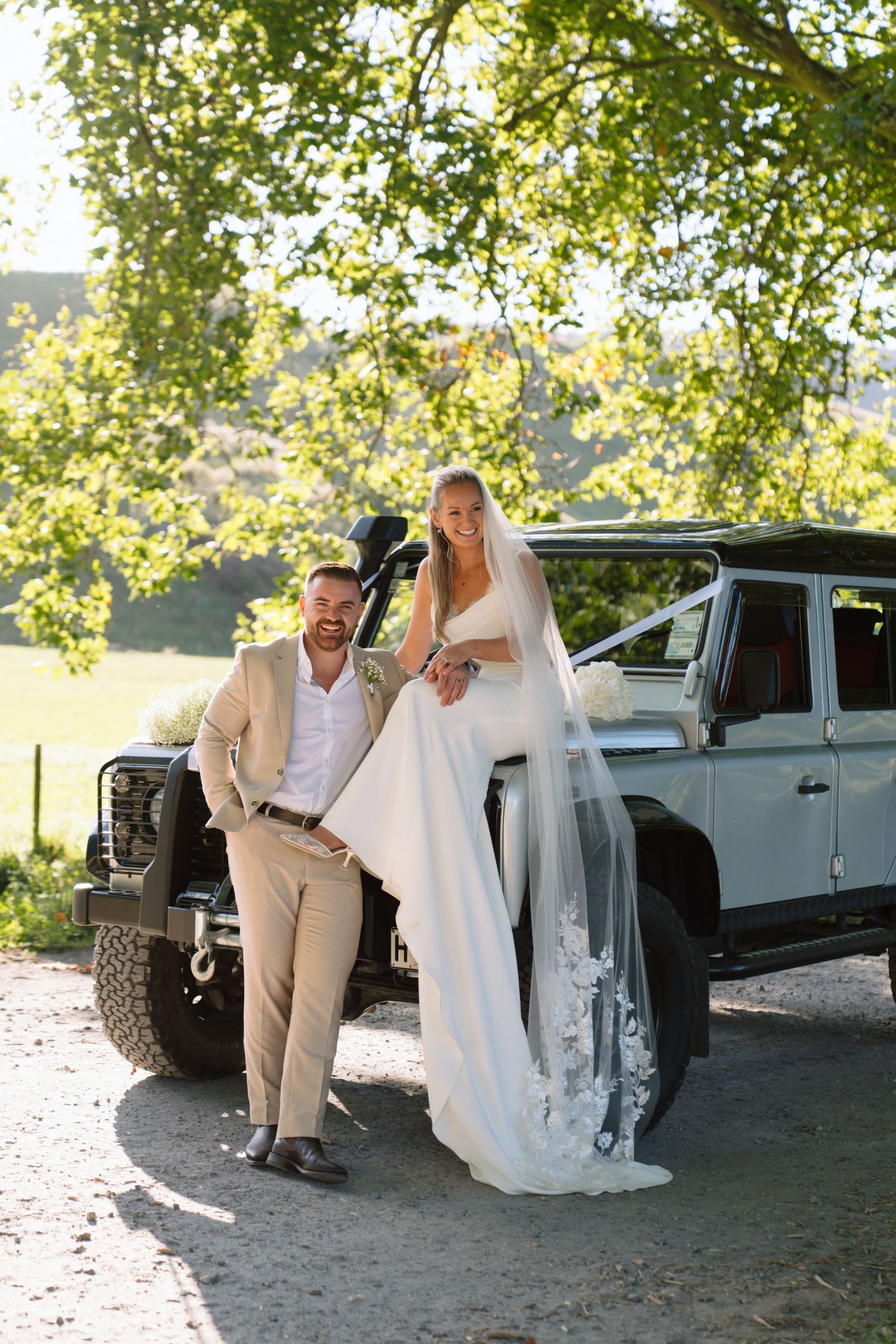 Bride and groom posing beside a vintage Land Rover Defender under green trees on their wedding day.