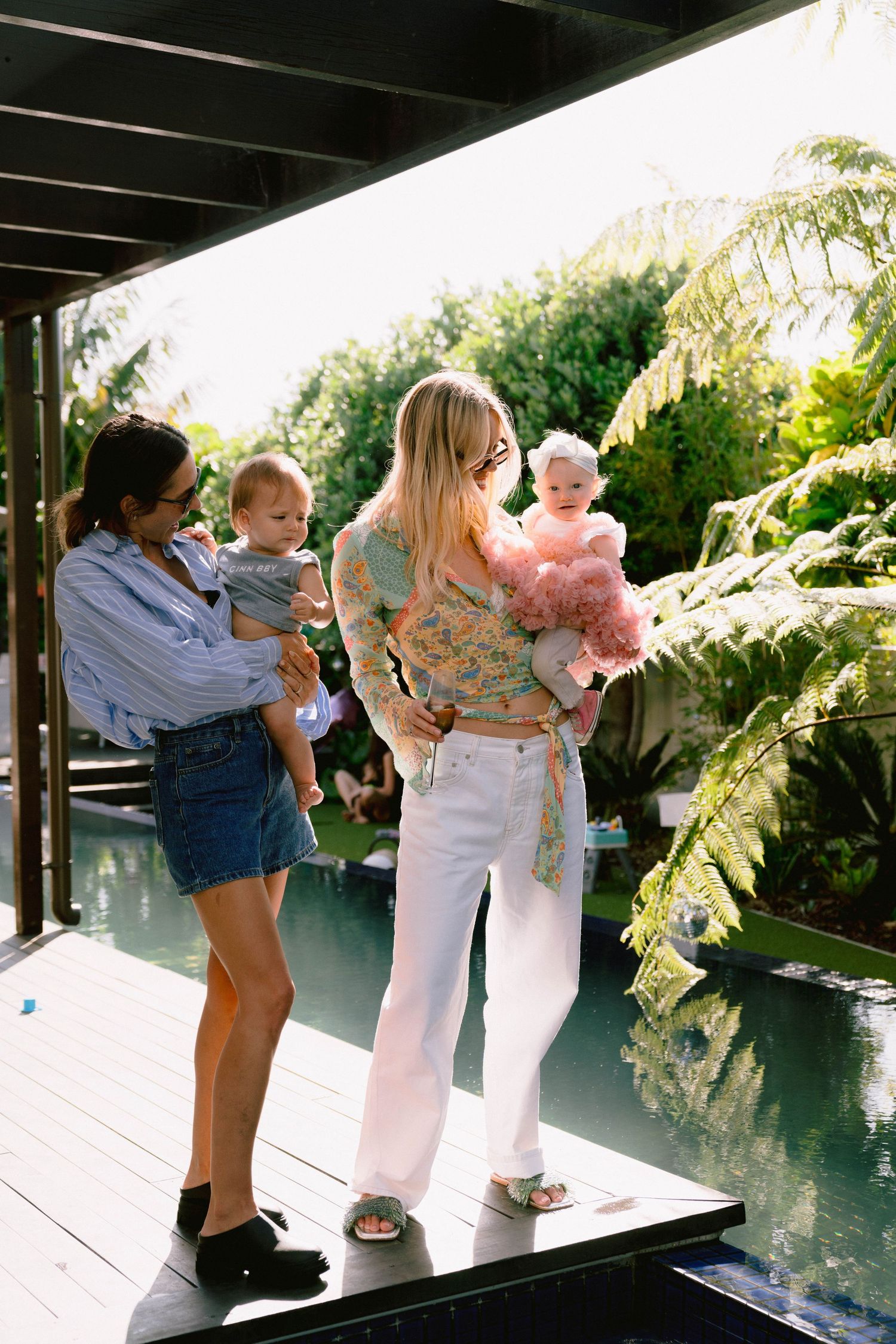 Friends gathered on a sunny deck near a pool surrounded by tropical ferns and greenery.