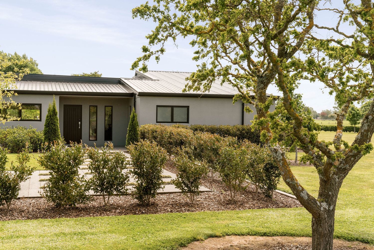Modern white house with black trim sits nestled among landscaped gardens and a mature tree in rural setting.