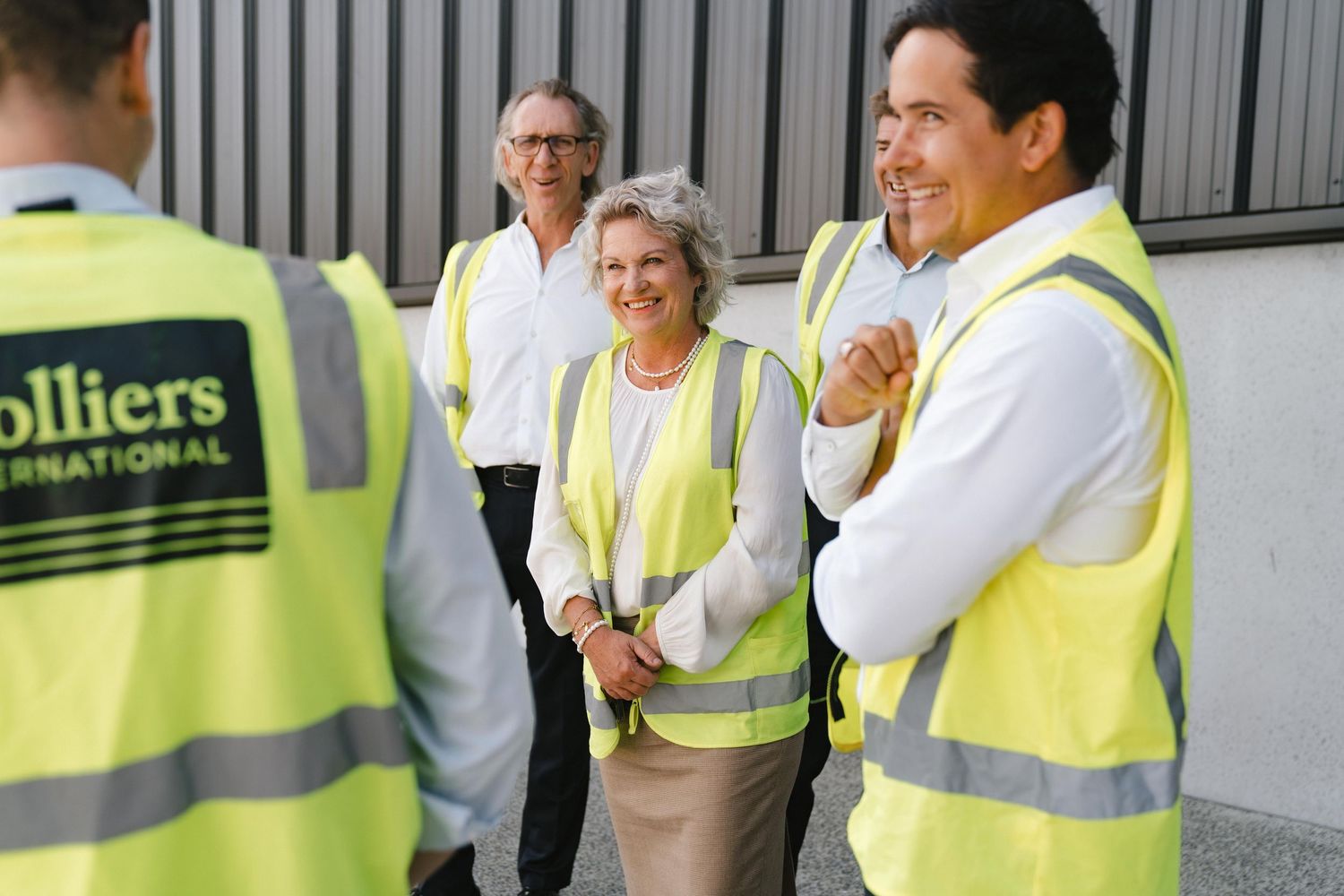 Group wearing Colliers International safety vests having discussion outside industrial building.