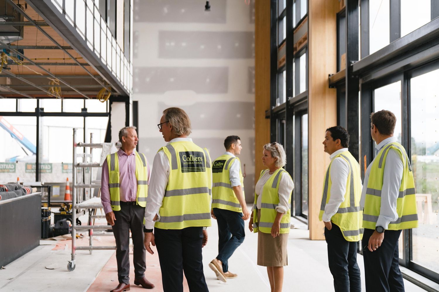 Team of professionals in safety vests meeting inside an unfinished commercial space with large windows.