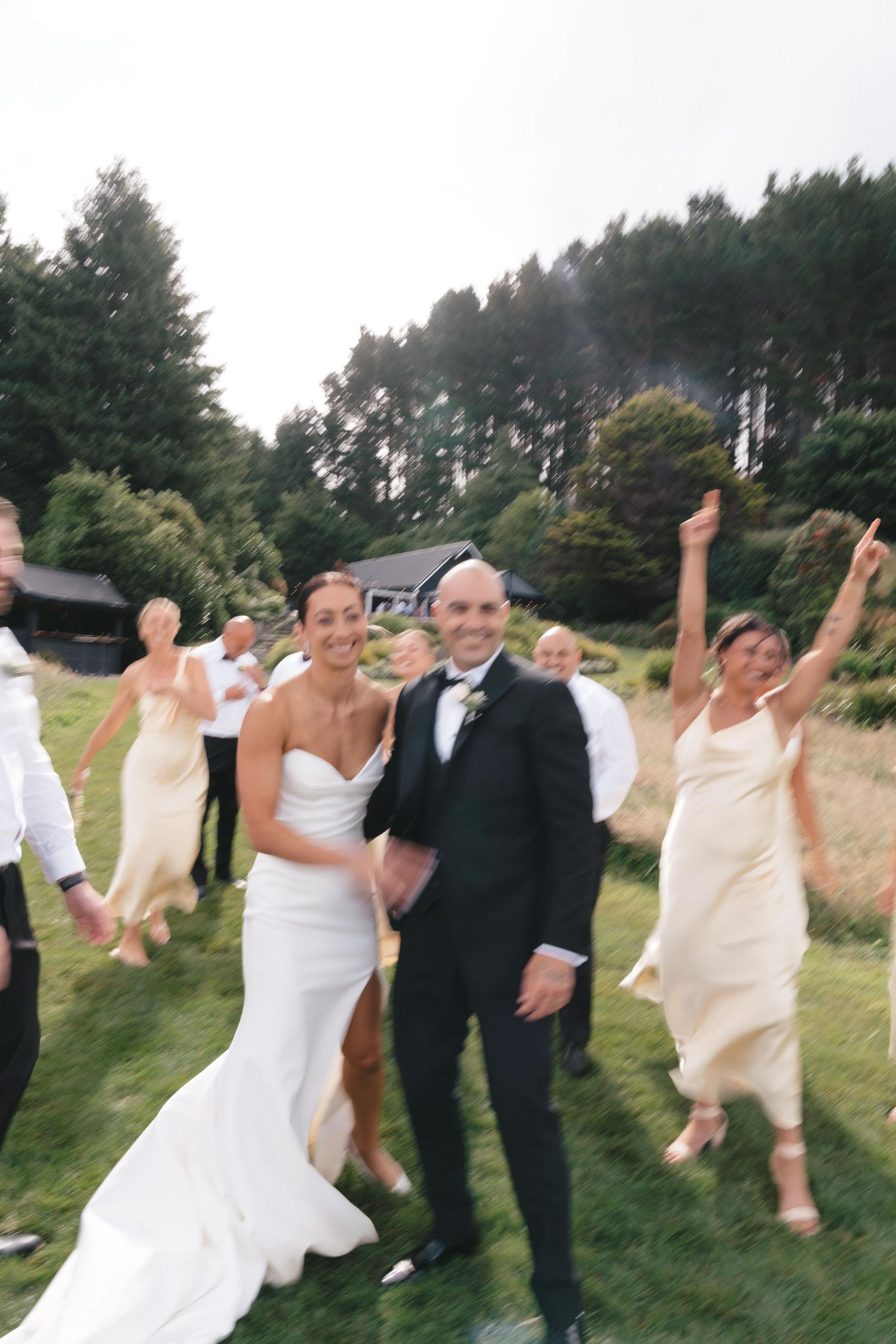 A wedding party celebrates joyfully on a grassy lawn with tall pine trees in the background during a summer afternoon.