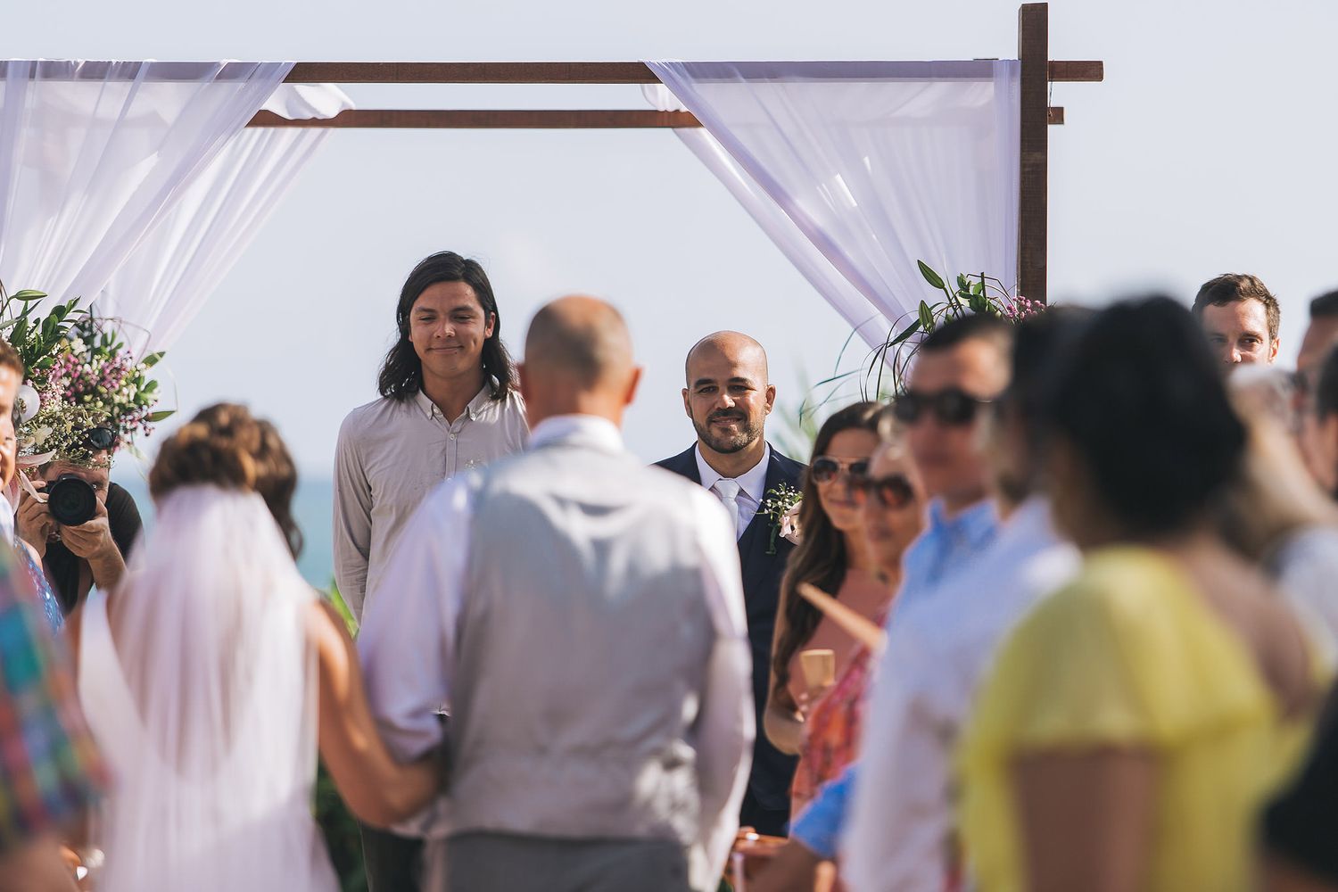 Outdoor wedding ceremony in progress under a white fabric arch.
