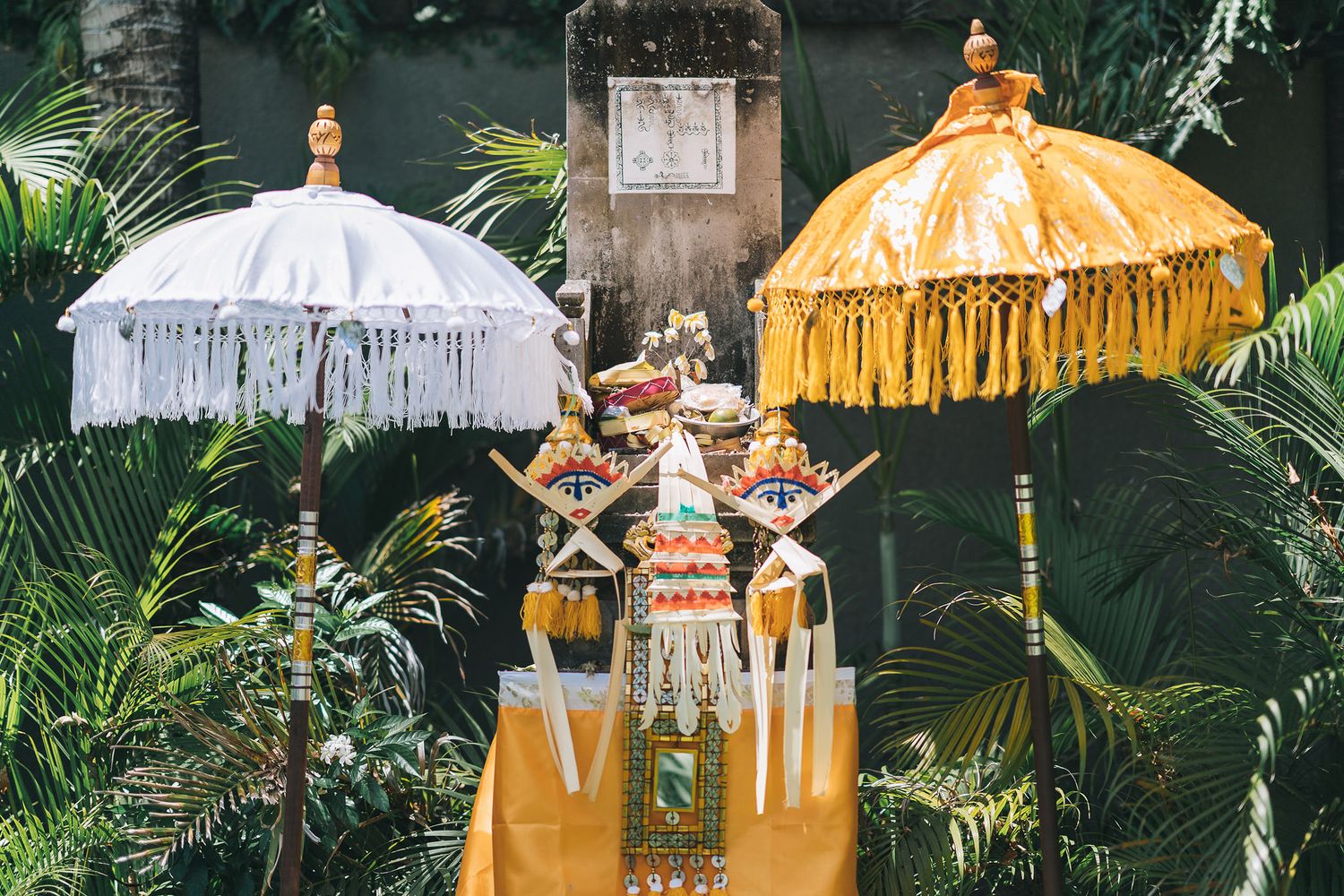 Two decorative Balinese parasols in white and yellow stand against tropical foliage in an outdoor garden setting.