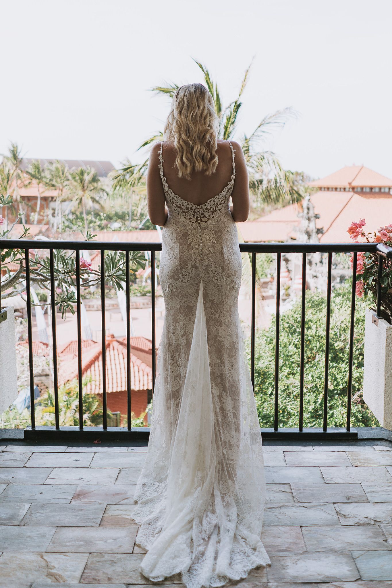 Back view of a bride in a lace wedding gown standing on a tropical balcony.