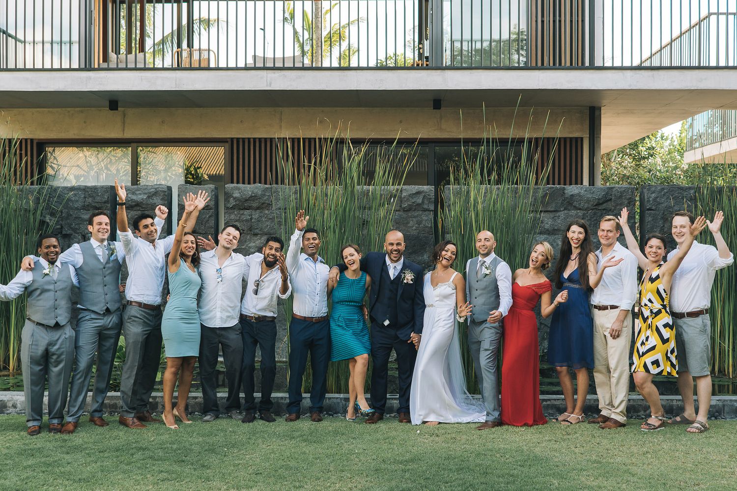 Wedding party poses for group photo on lawn with modern architecture in background.