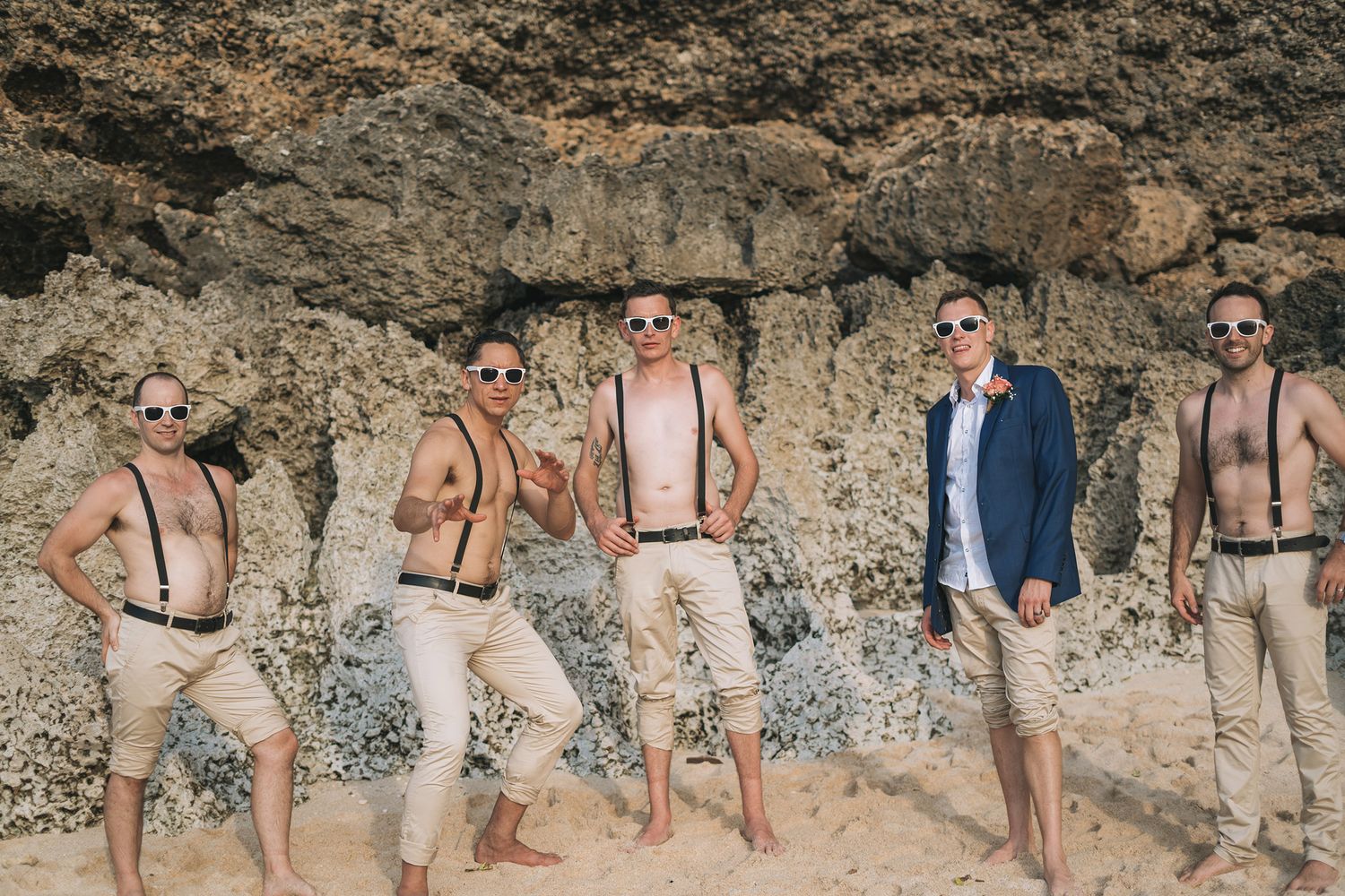 Group of groomsmen in matching attire posing against rocky cliff wall wearing sunglasses.
