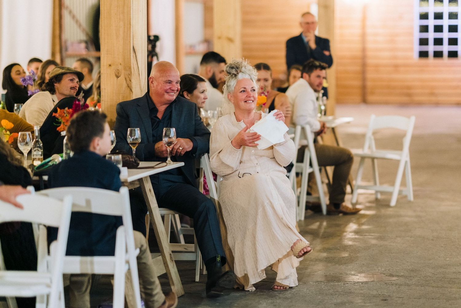 Wedding celebration in rustic venue with guests seated on white folding chairs.