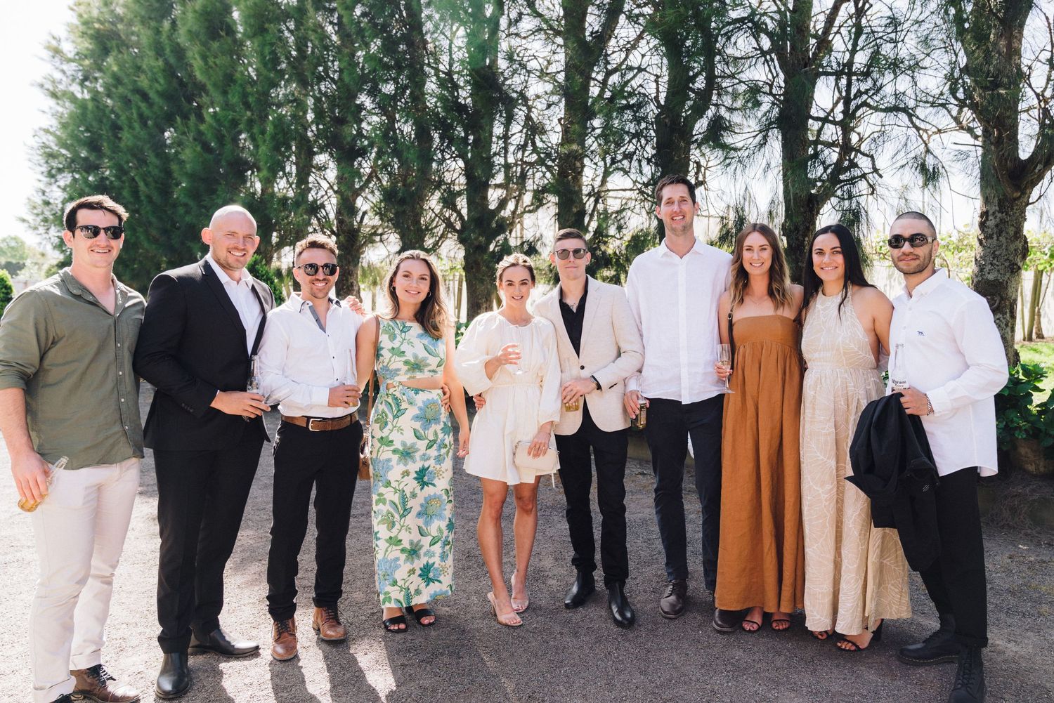 A group photo of wedding guests in casual attire standing outdoors against a row of trees.