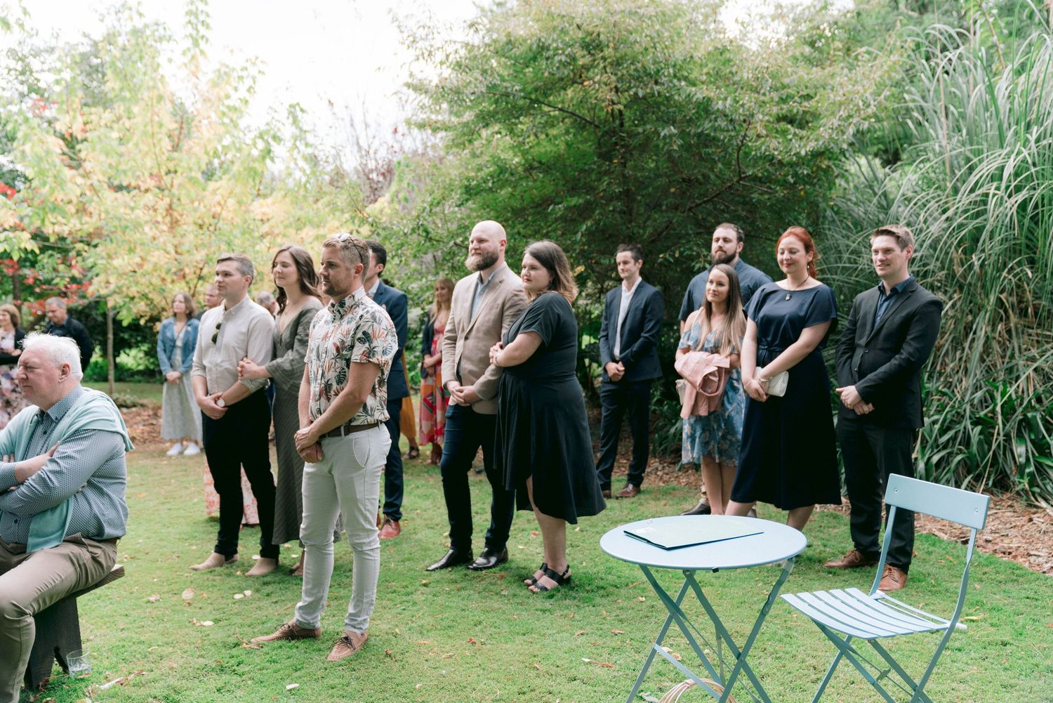 Wedding guests gather outdoors on a lawn near garden foliage for a ceremony.