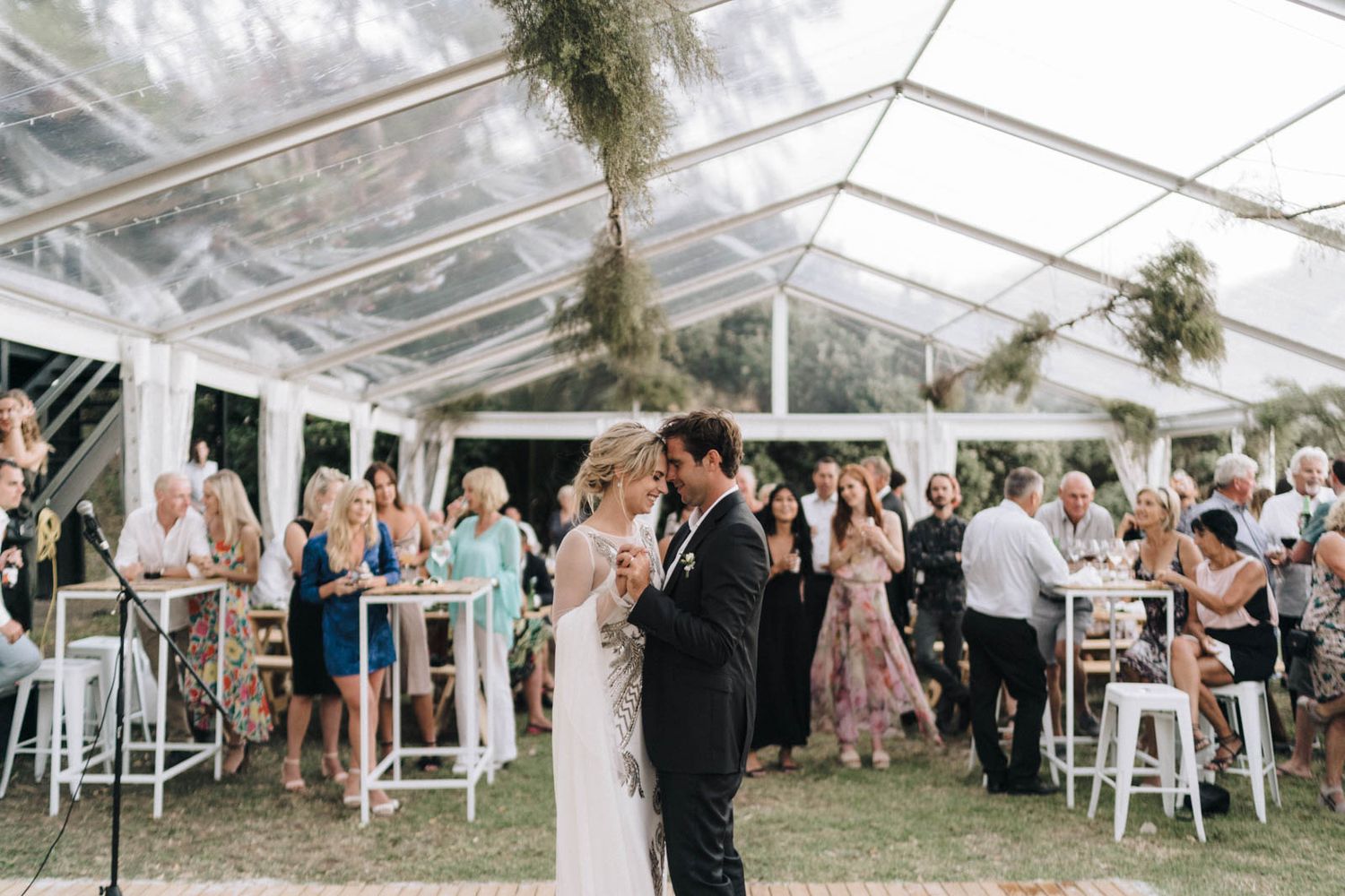 Wedding guests dance together under a white tent while string lights illuminate the outdoor reception venue.