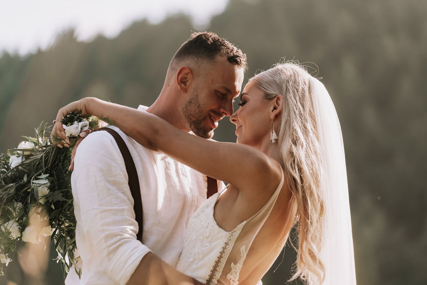 Two people in formal wedding attire embrace romantically in an outdoor setting with blurred greenery in the background.
