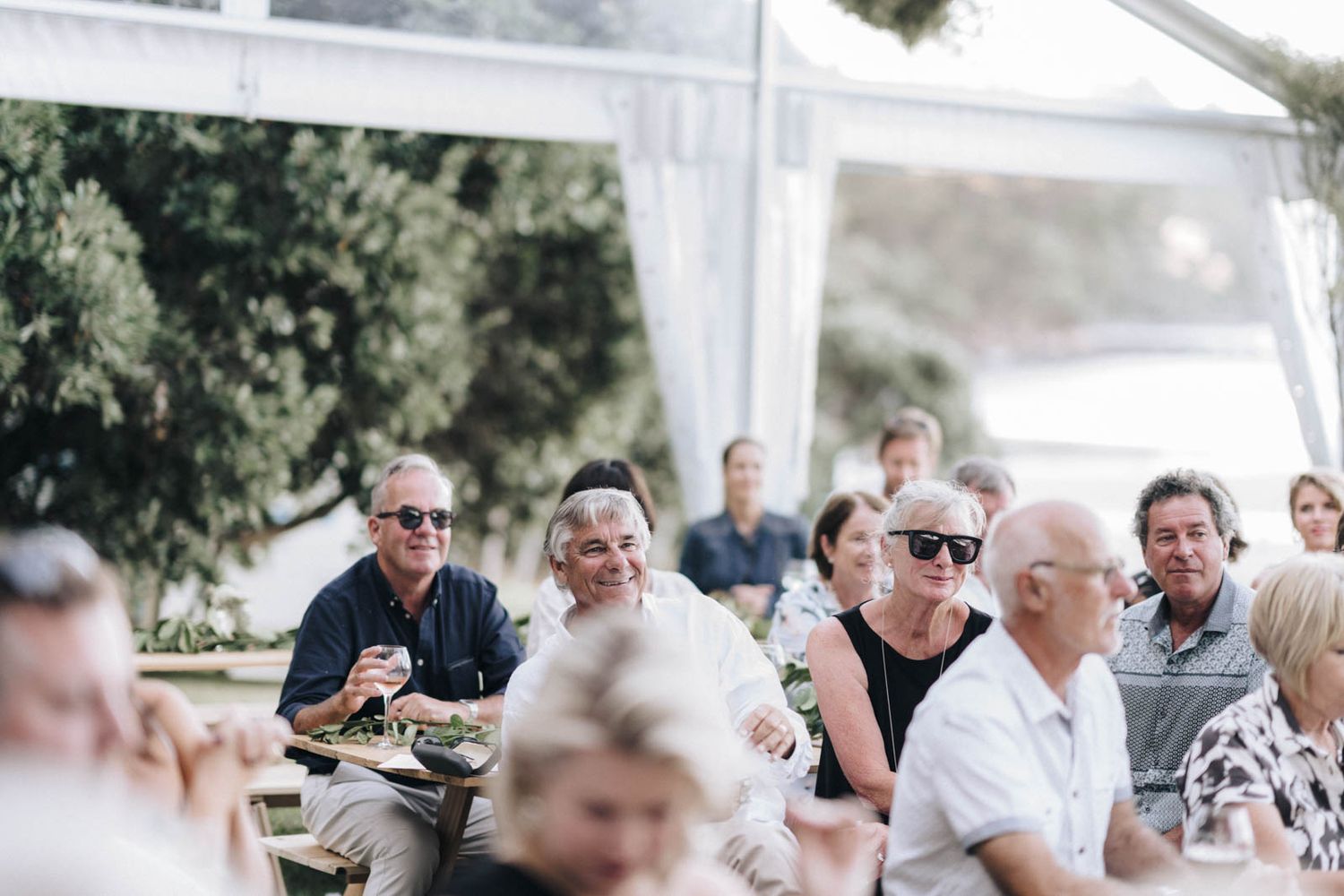 Wedding guests seated under a white tent canopy enjoying a casual outdoor reception with bright natural lighting.