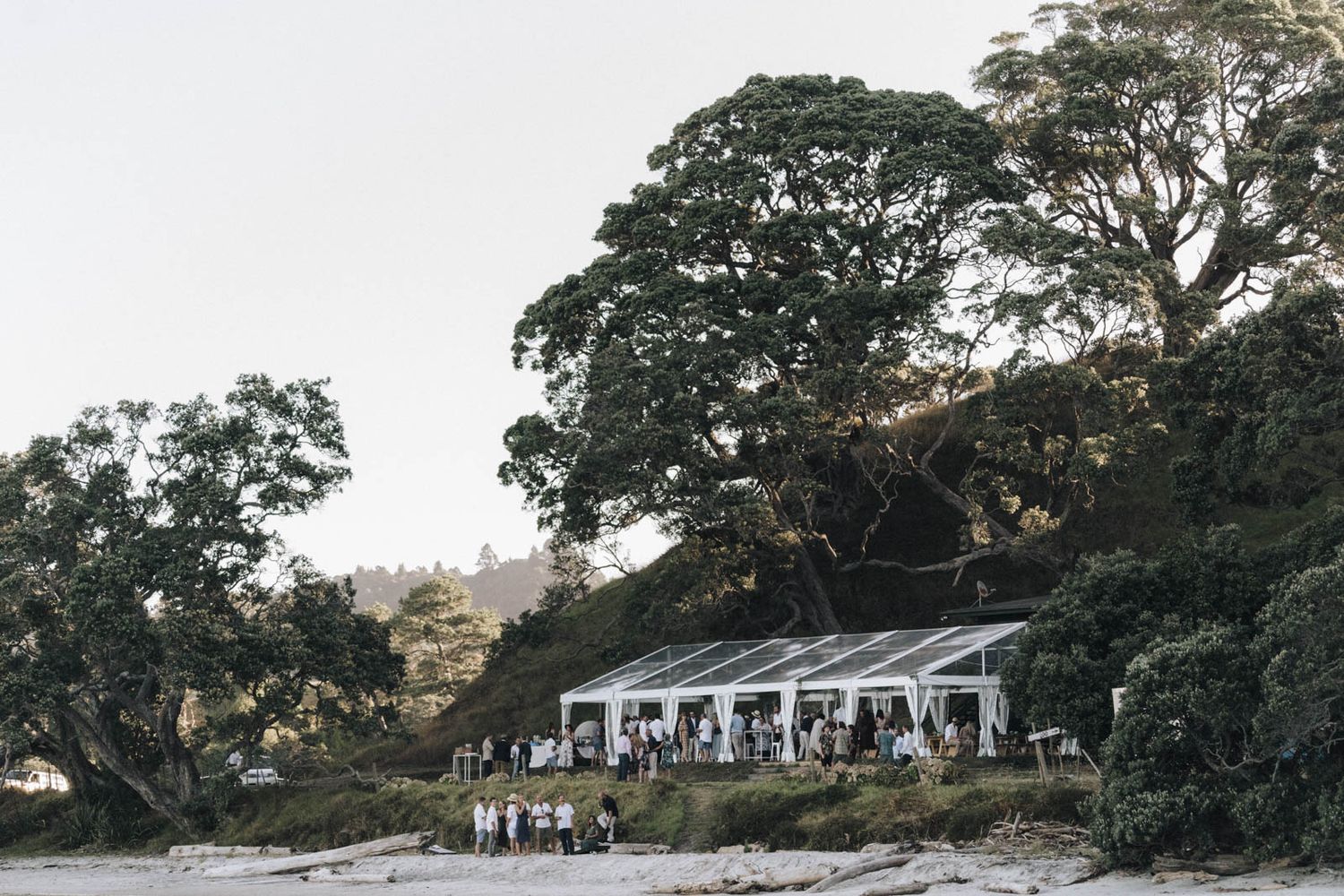 Elegant outdoor wedding reception tent set up along a waterfront beach with tall trees in the background at dusk.