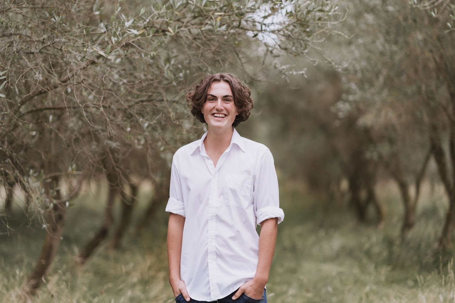 Young person in white button-up shirt smiling while standing in an olive grove with soft natural lighting.