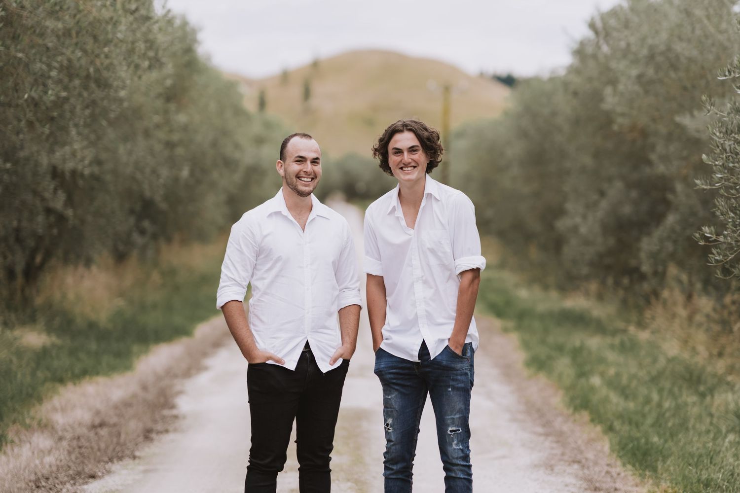 A couple in matching white shirts and dark pants walk together on a winding dirt road through olive trees.