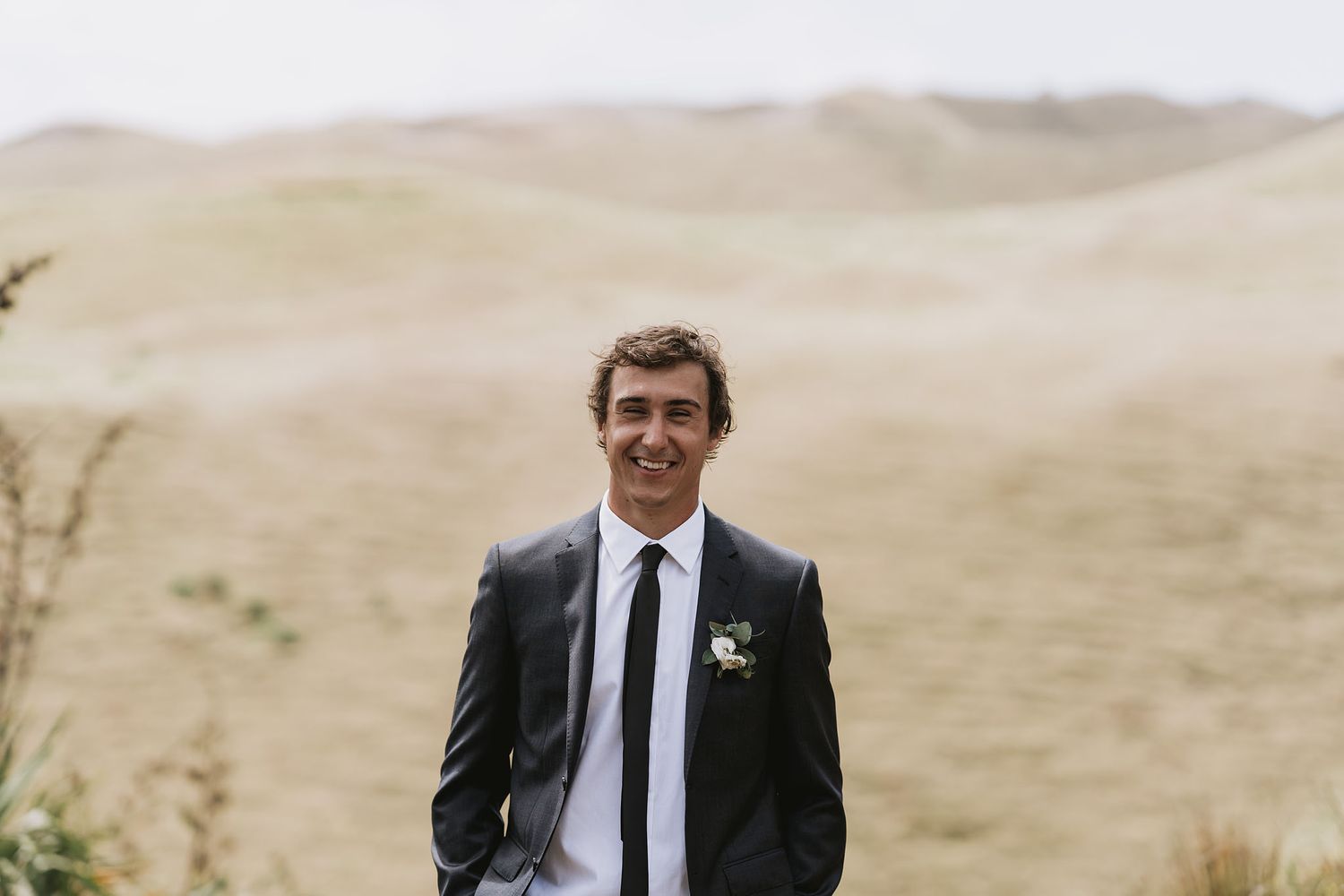 Man in a black suit stands in a desert landscape with rolling sand dunes in the background.