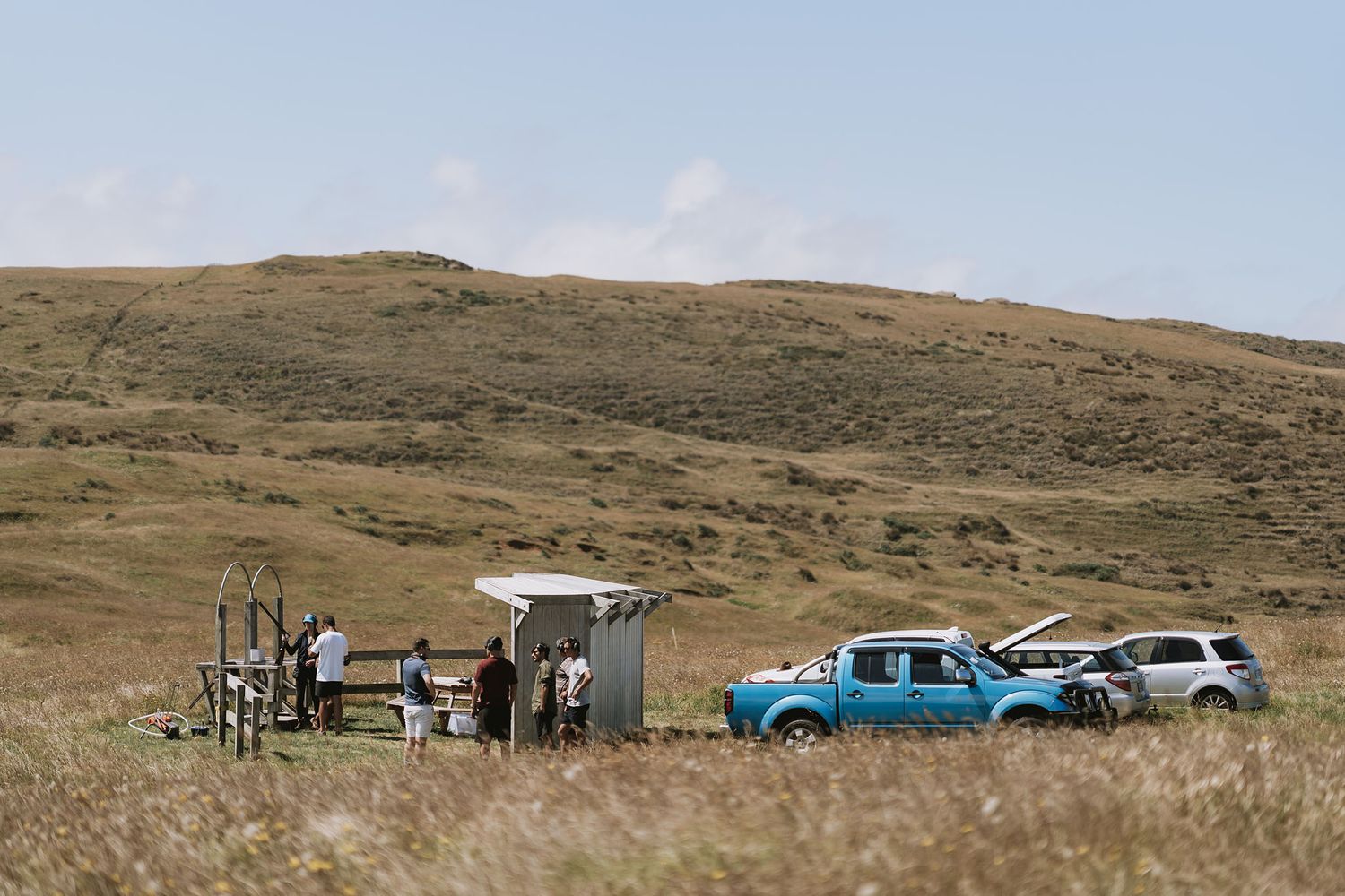Two vehicles parked near a camping setup with equipment and people in a vast grassland landscape under blue skies.