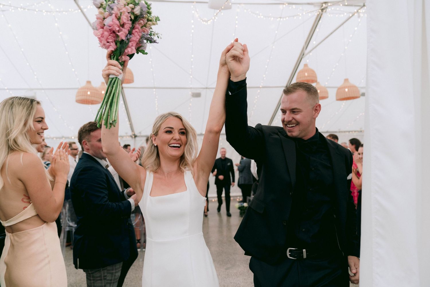 Wedding guests dance and laugh during a joyful reception celebration under white tent lighting.