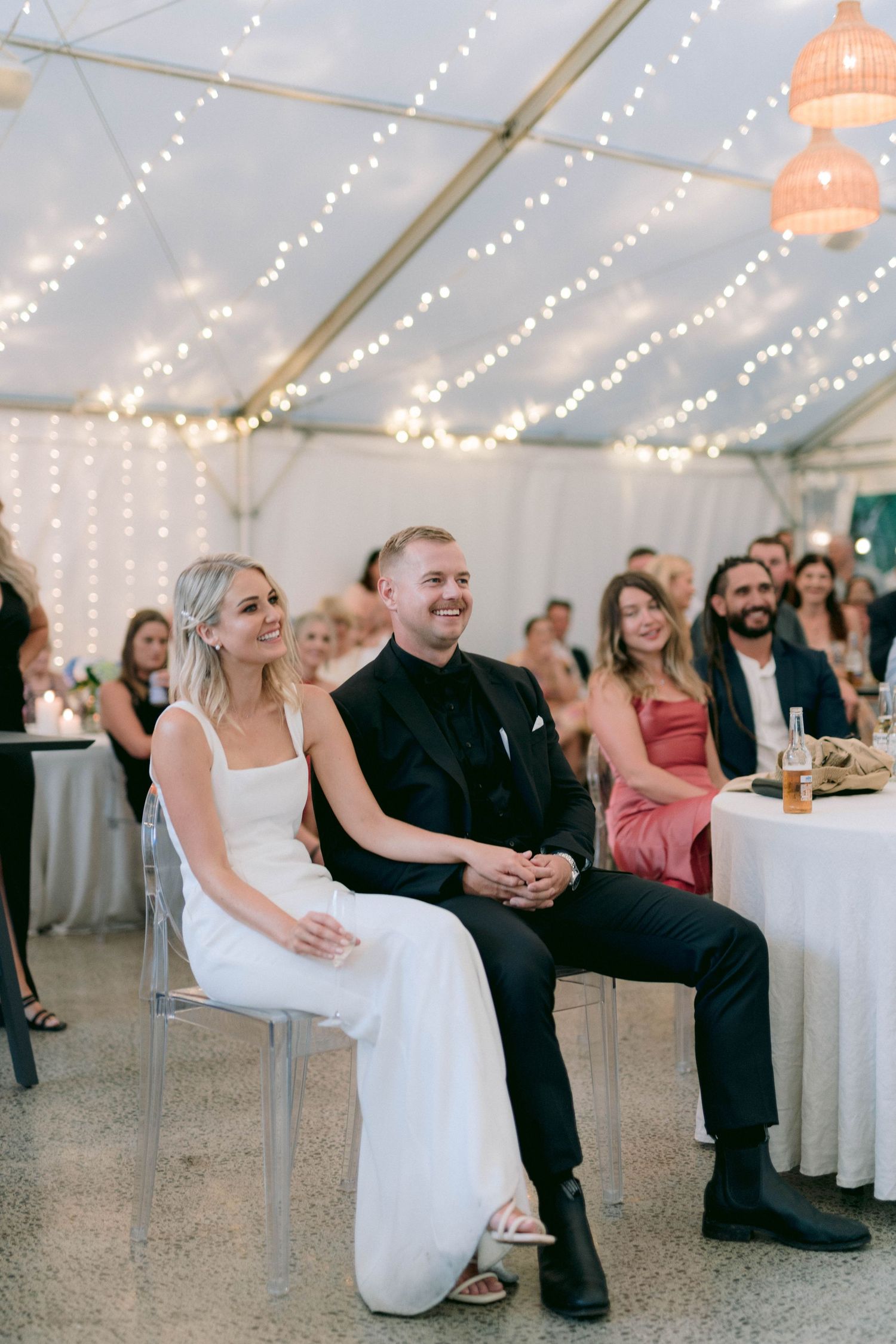 Wedding guests enjoy a reception under a tent decorated with string lights and peach lanterns.
