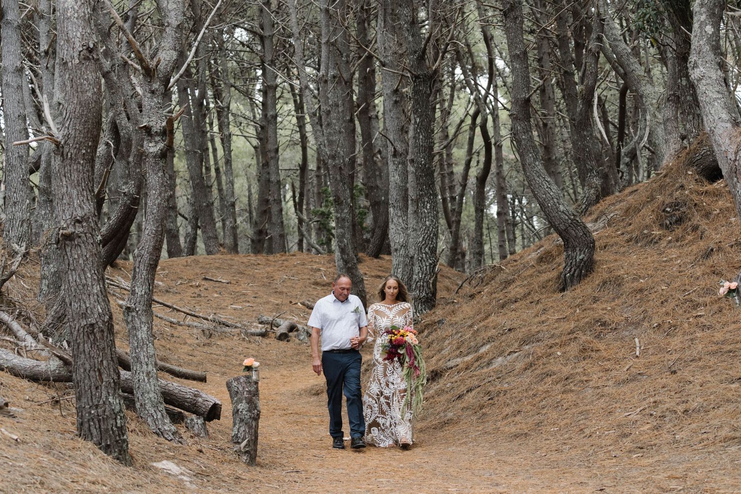 Two people walk hand in hand along a dirt path through a grove of tall birch trees on a sloping hillside.