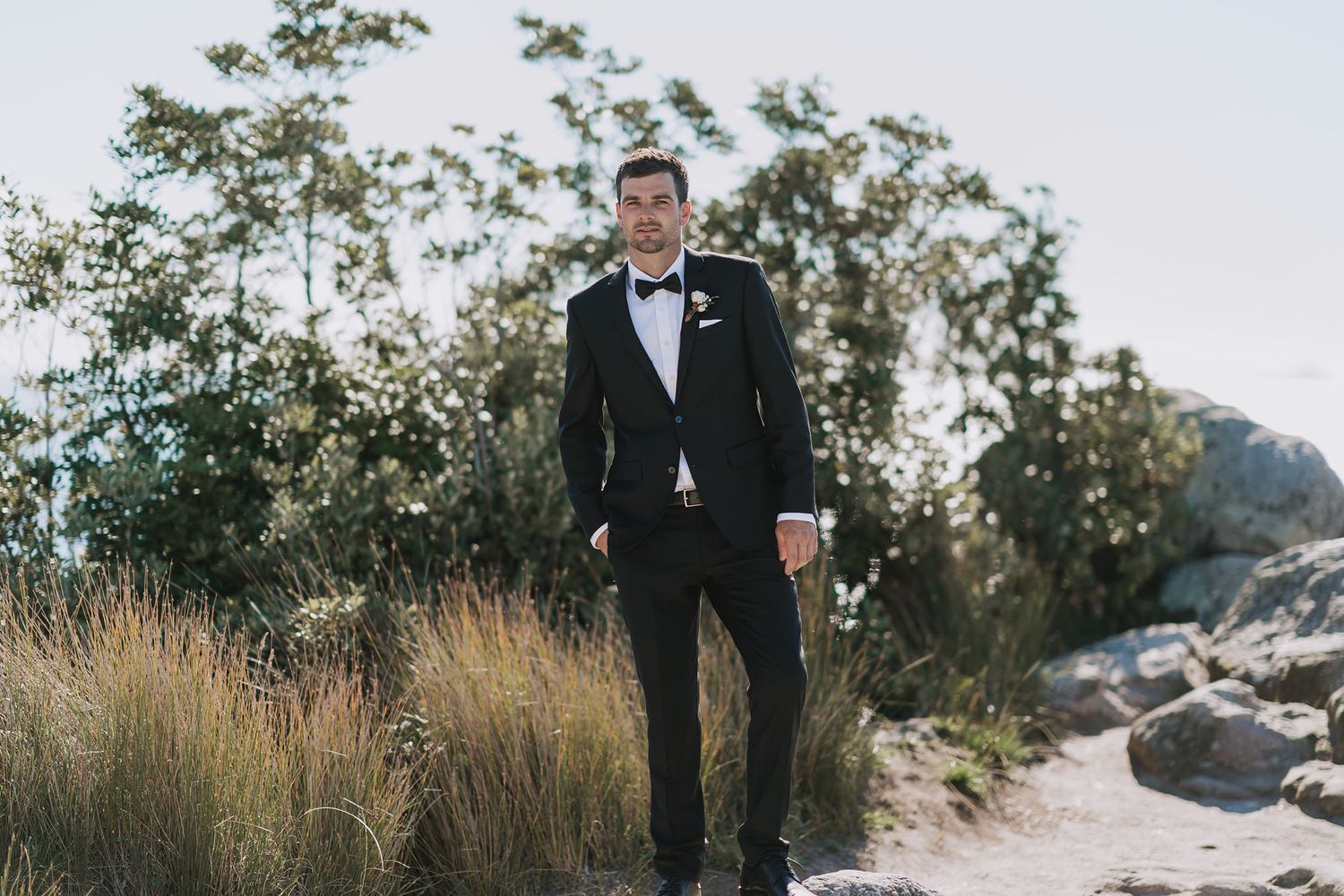A person in a classic black tuxedo and bow tie stands amongst tall grasses near rocky terrain during daytime.