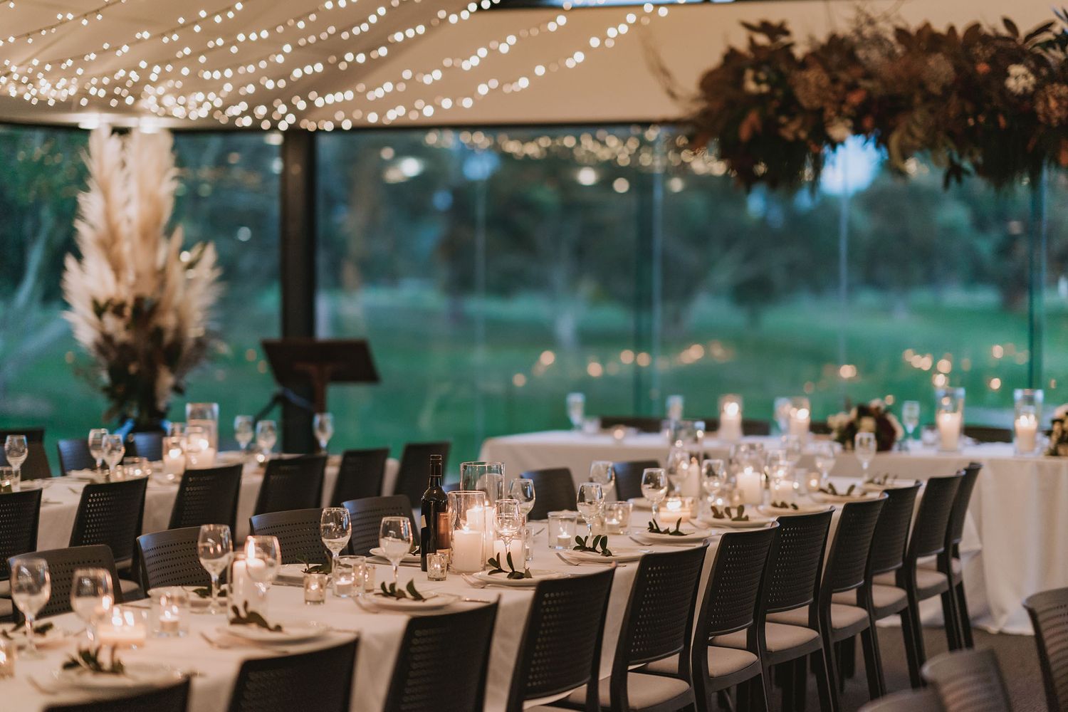 Elegant wedding reception tables set with white linens, glassware and string lights under a covered outdoor venue.