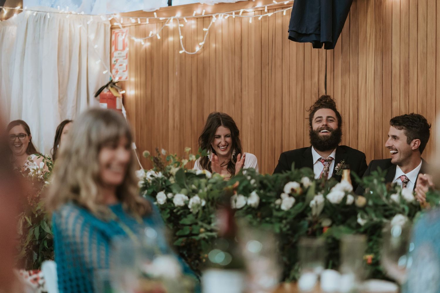 Wedding guests share a lighthearted moment at a reception decorated with string lights and greenery.