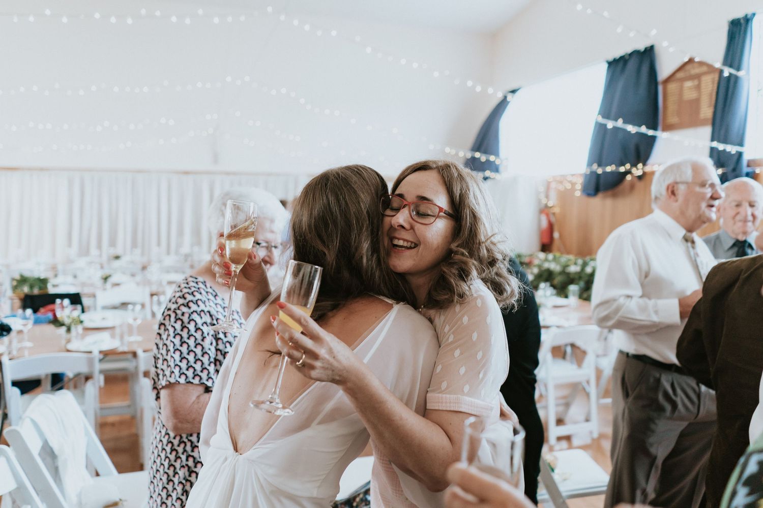 Two people embrace and smile while celebrating at a bright indoor wedding reception with champagne glasses.