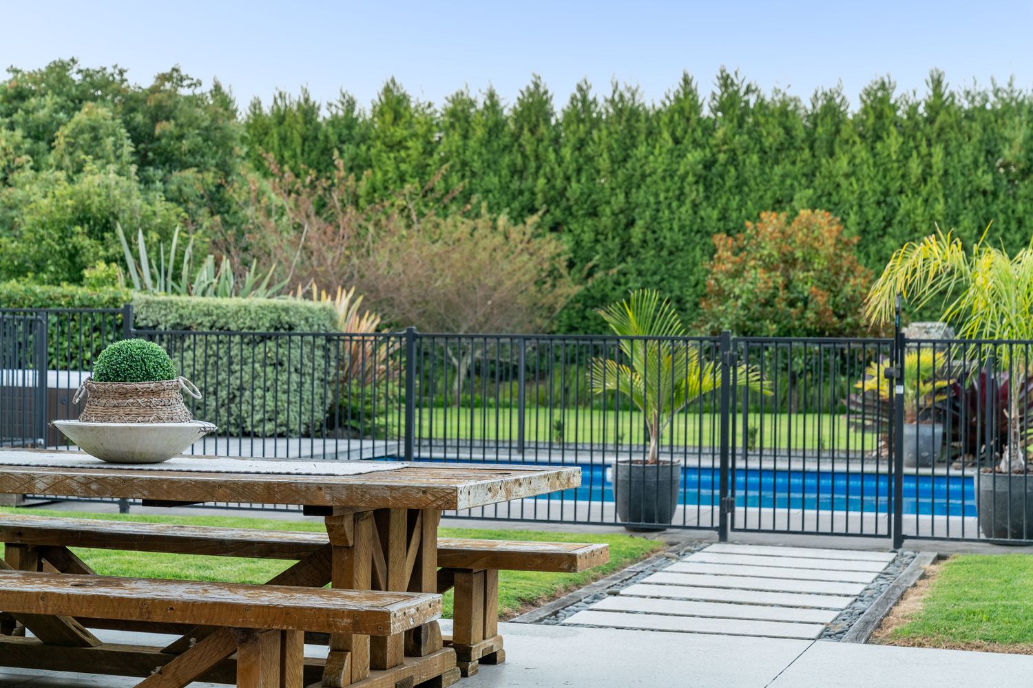 Backyard view of swimming pool with safety fence and wooden picnic table in foreground.