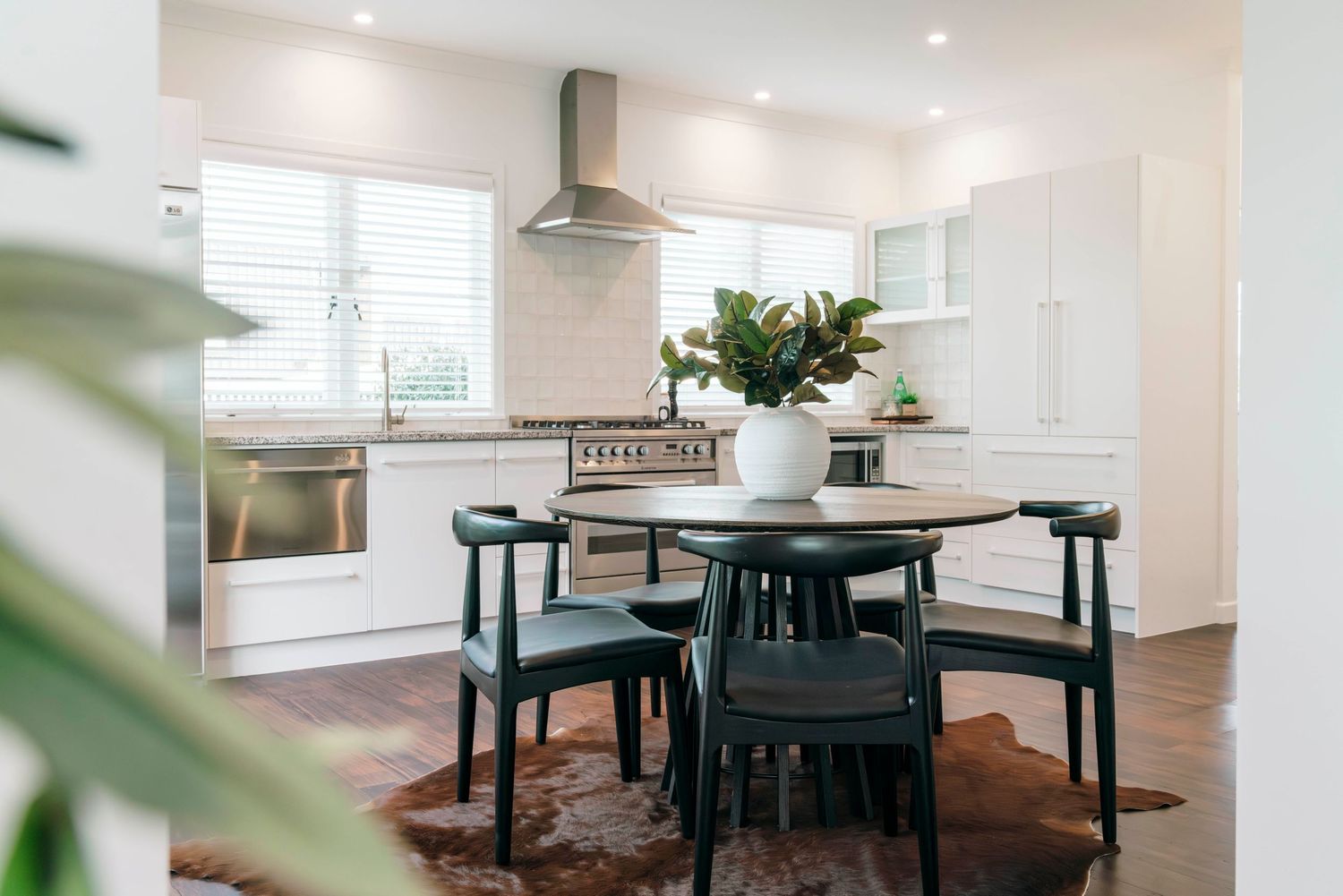 Modern white kitchen with dark dining set, stainless steel hood, and hardwood floors.